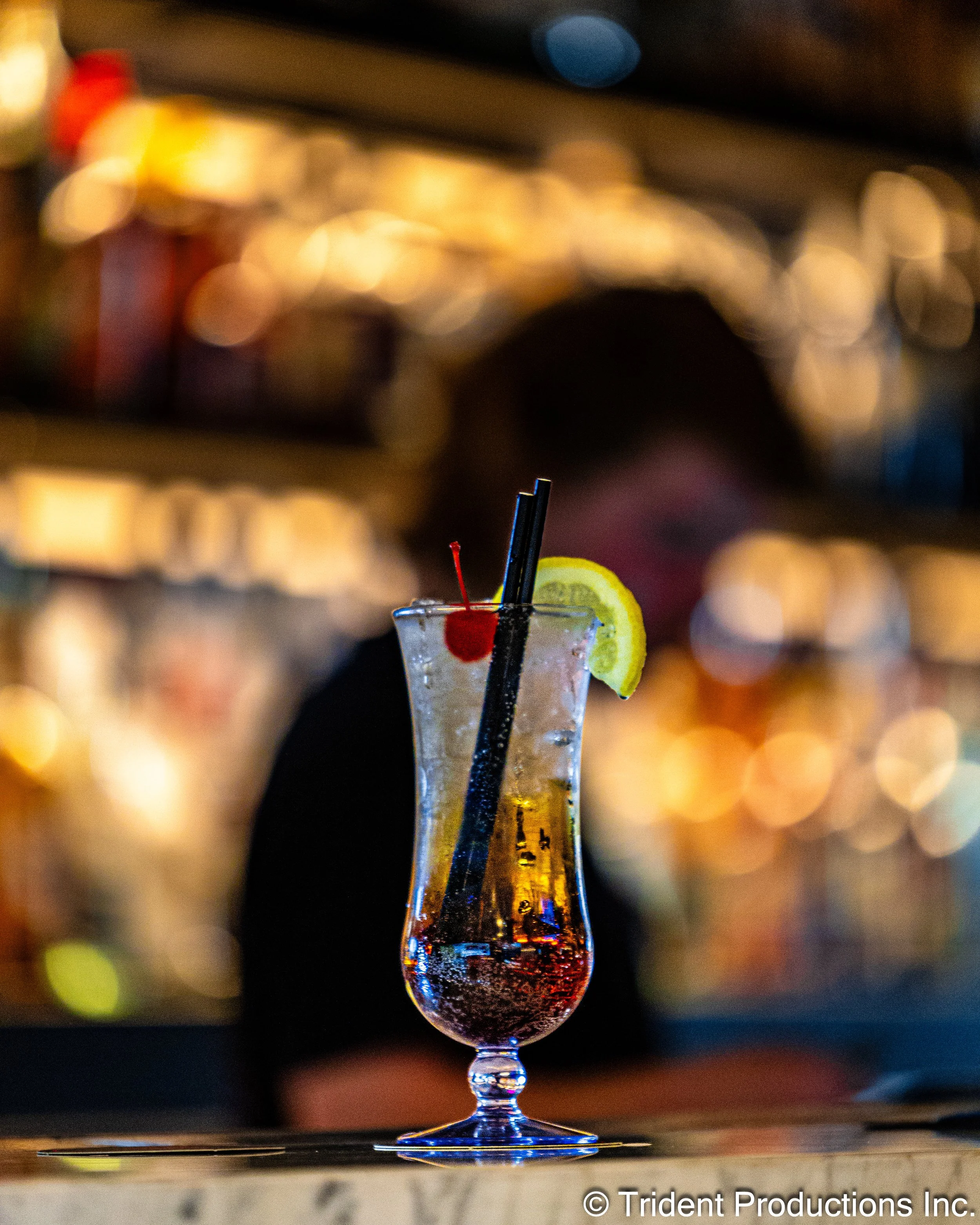 A tropical cocktail garnished with a lemon wedge and a maraschino cherry, placed on a bar counter with a blurred background of a bar or restaurant.