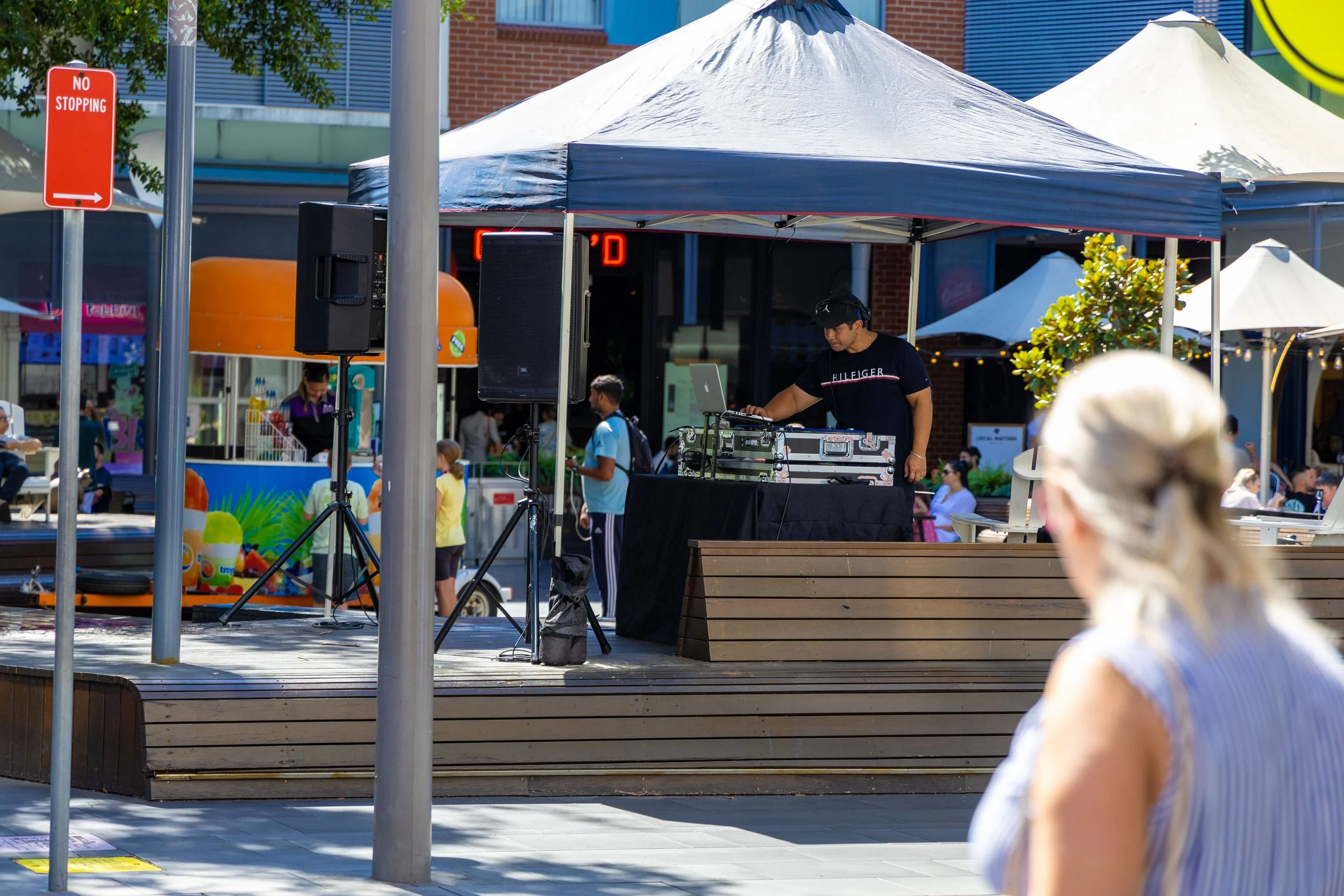 DJ performing music at an outdoor event with a crowd of people in the background, a vendor stand with ice cream and other treats, and white tents.