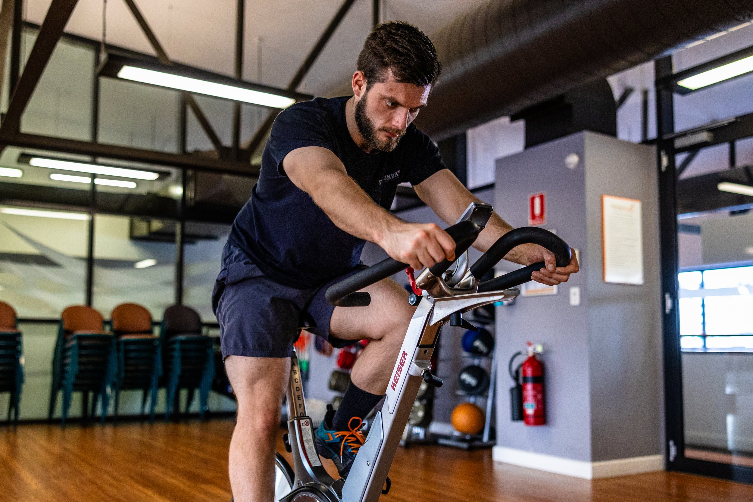 A man with a beard and short dark hair is exercising on a stationary bike indoors, wearing a black t-shirt and shorts.