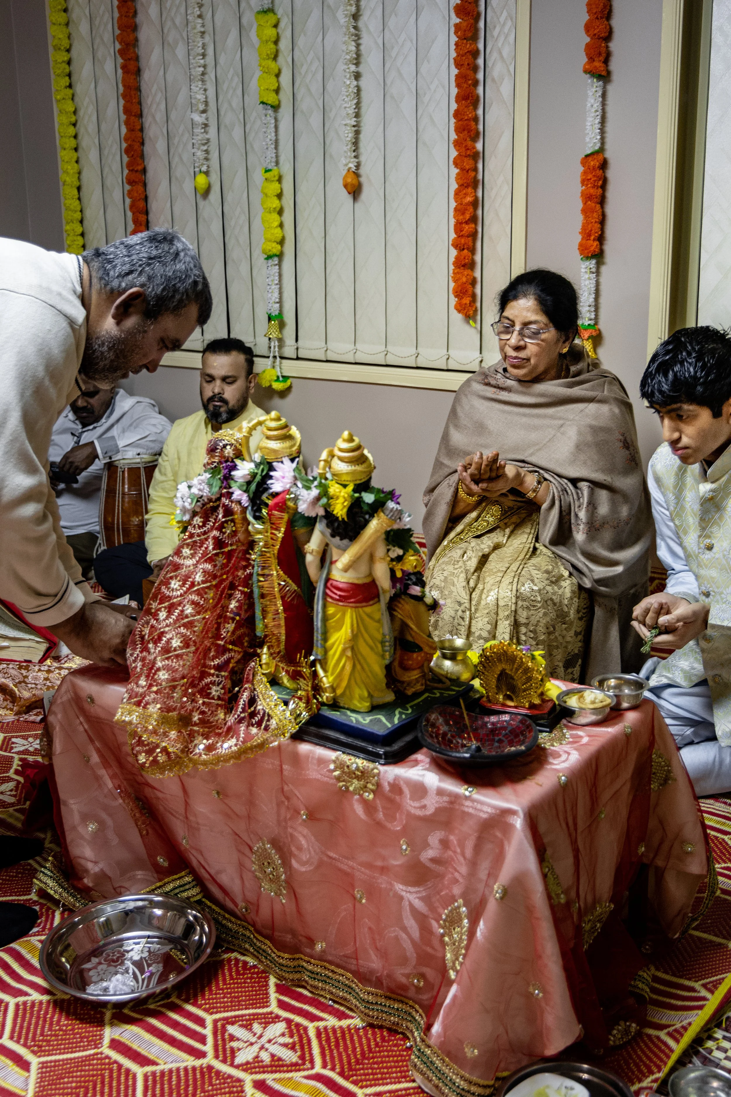 Indian family performing a religious ritual around a decorated table with various idols and offerings, with decorative flowers hanging on the wall behind them.