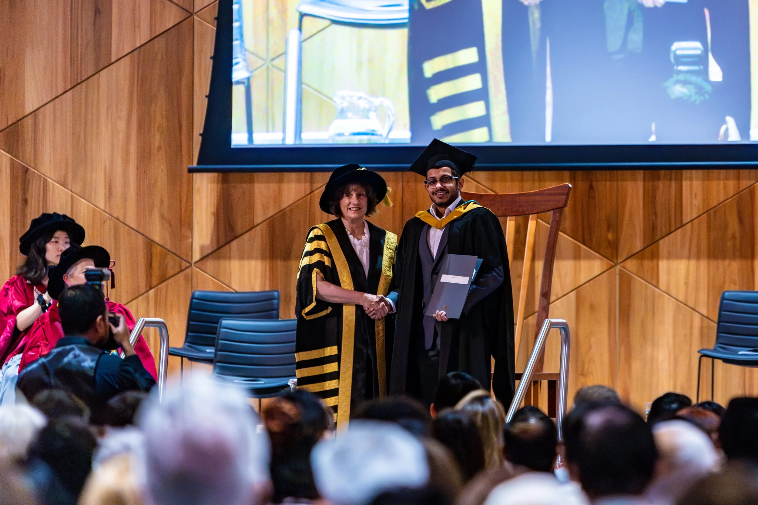 A graduation ceremony where a graduate in cap and gown shakes hands with a faculty member in academic regalia on stage, while photographers and an audience observe.