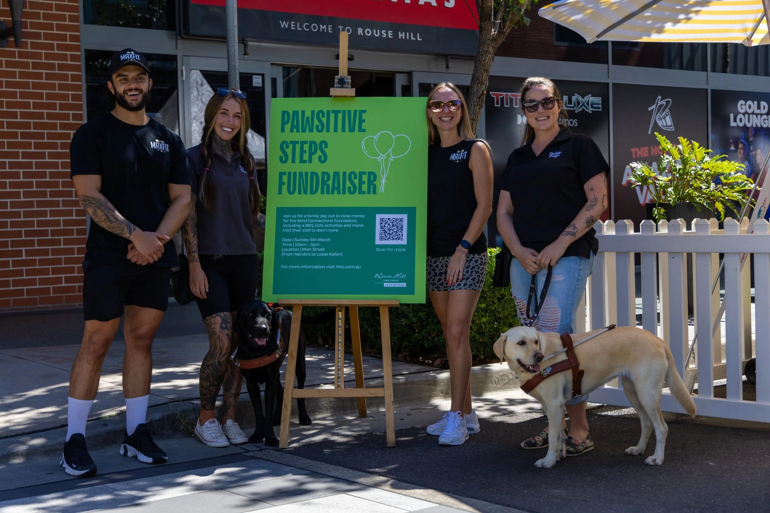 Four people standing outdoors in front of a bright green sign that reads 'Pawsitive Steps Fundraiser,' with two dogs on leashes and a yellow striped umbrella and storefront in the background.