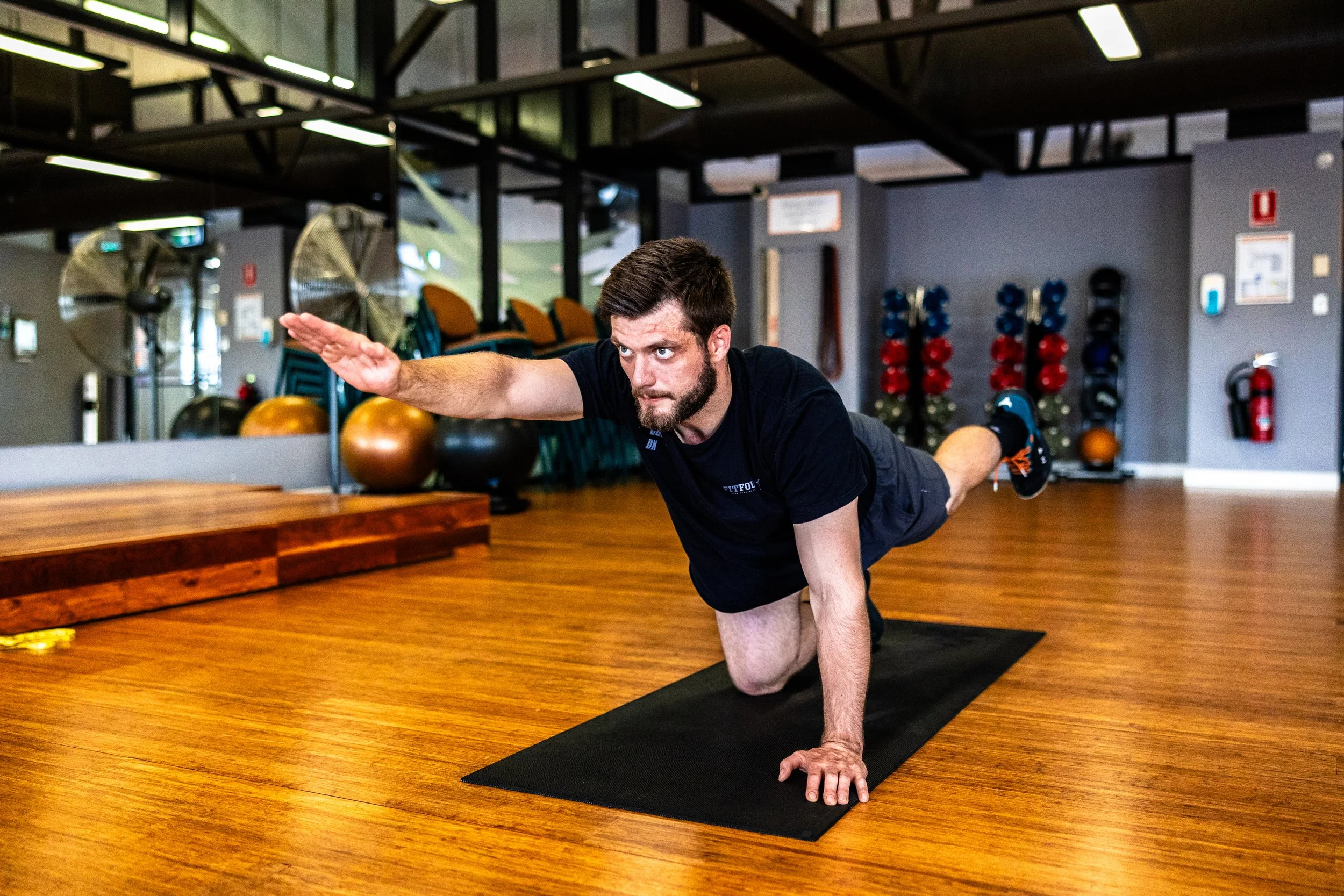 Man performing a yoga pose on a black mat in a gym with exercise equipment and wooden flooring.