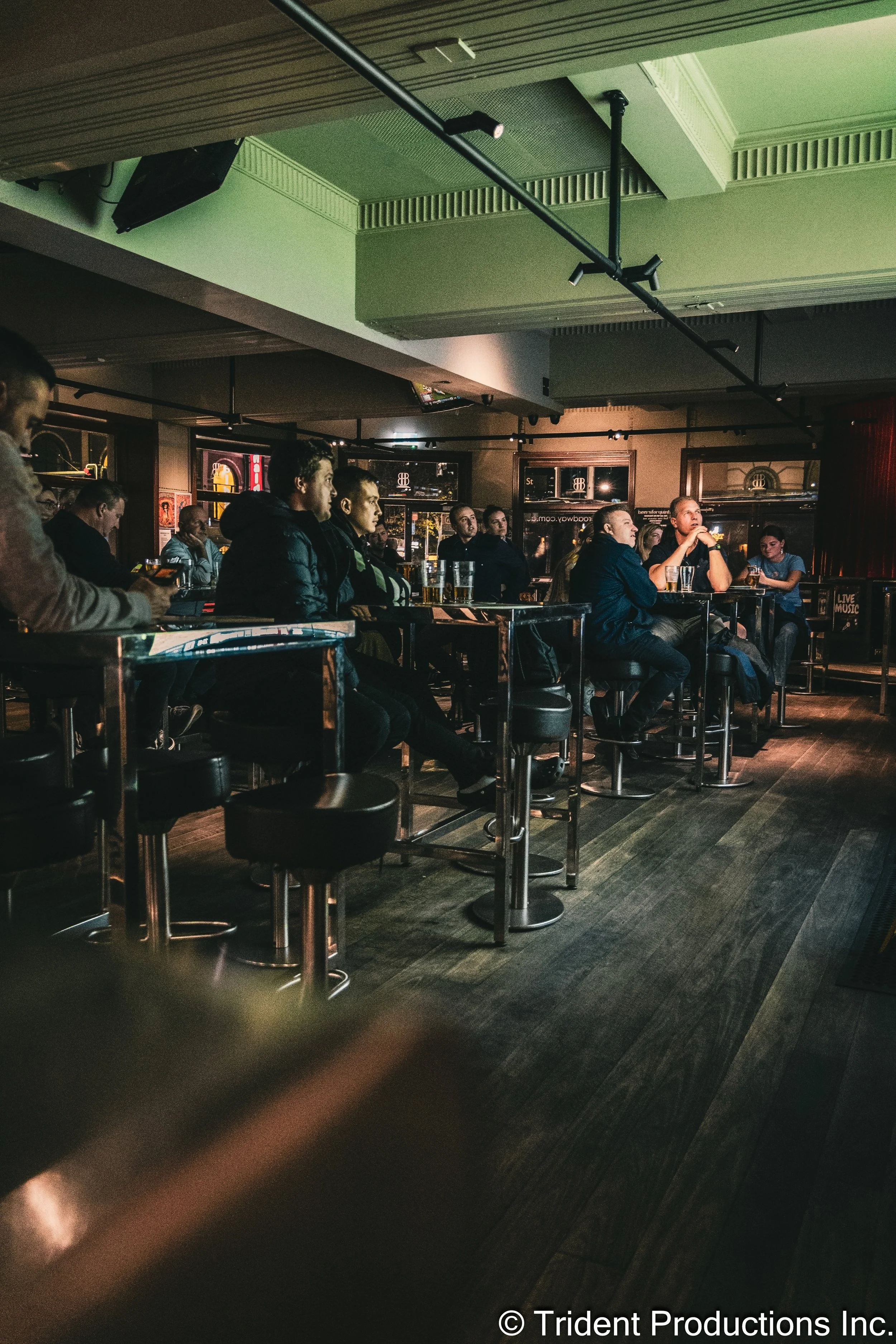 Audience watching a performance at a bar or club, seated at high tables with drinks in front of them, illuminated by stage lighting with a dark interior.