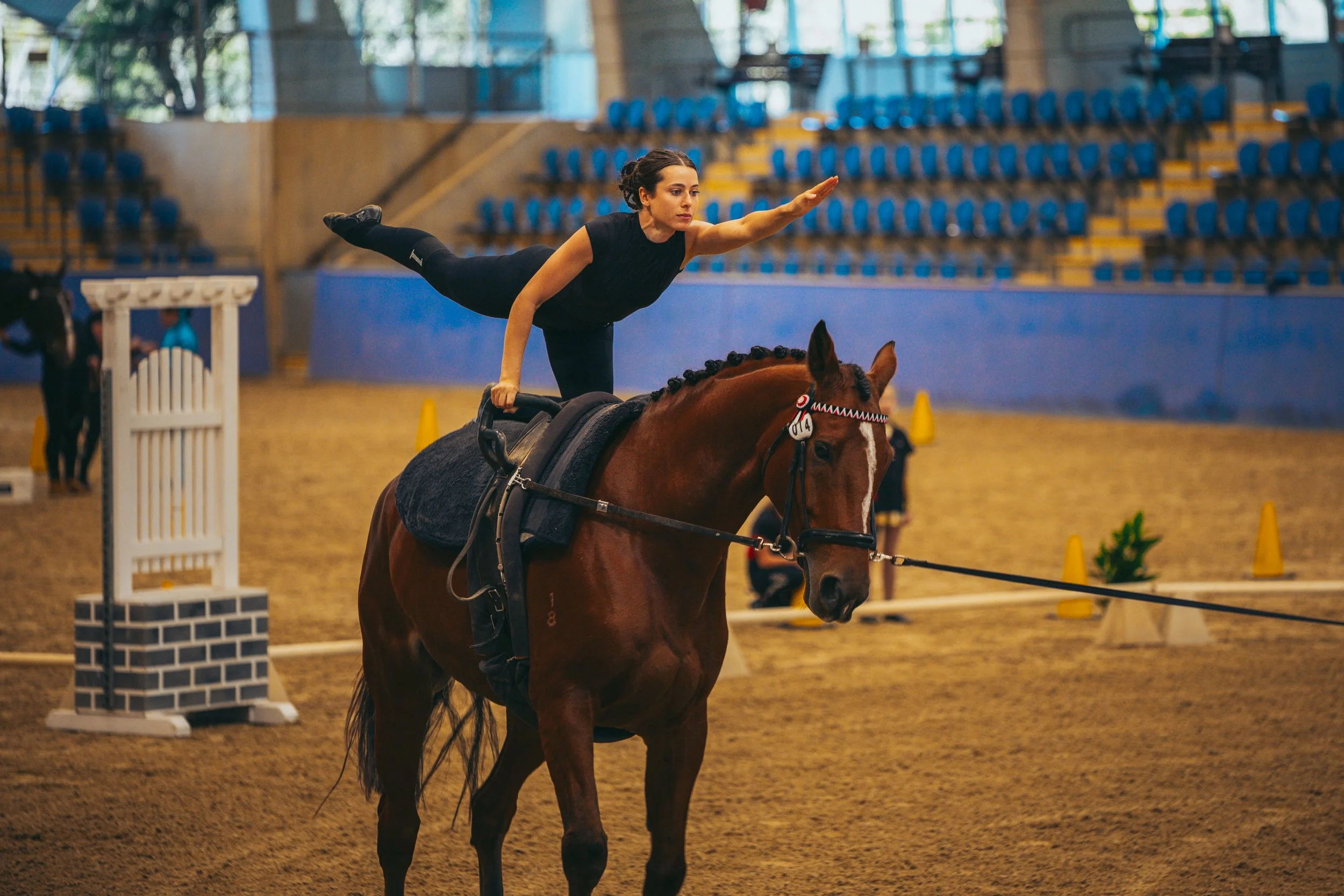 A woman performing a balancing act on a brown horse inside an indoor equestrian arena. She is extending her right arm forward and leaning to the side while holding onto the horse. The arena has blue seating and yellow cones in the background.
