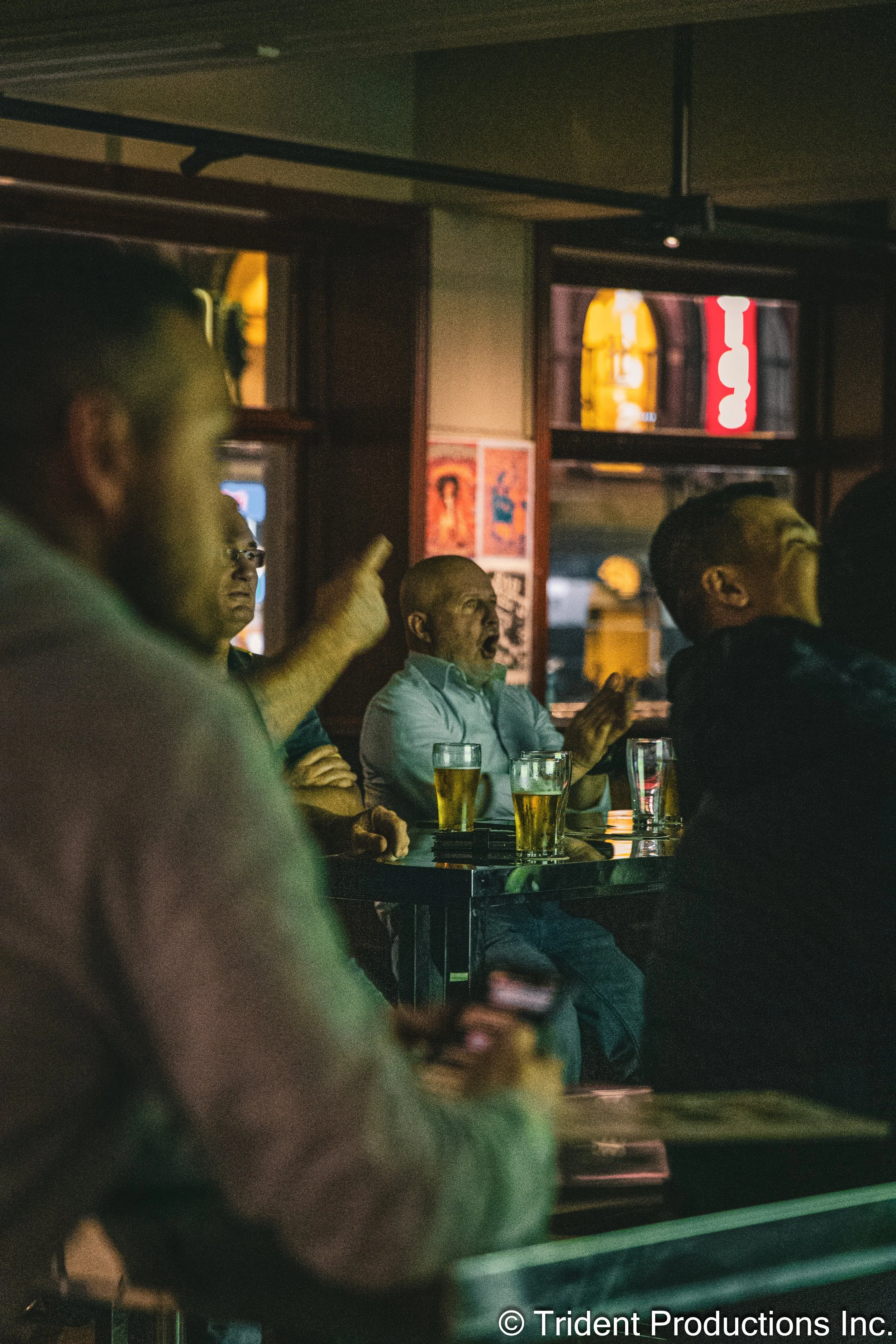 A man sitting at a bar or pub, engaged in an animated conversation, with glasses of beer on the table in front of him.