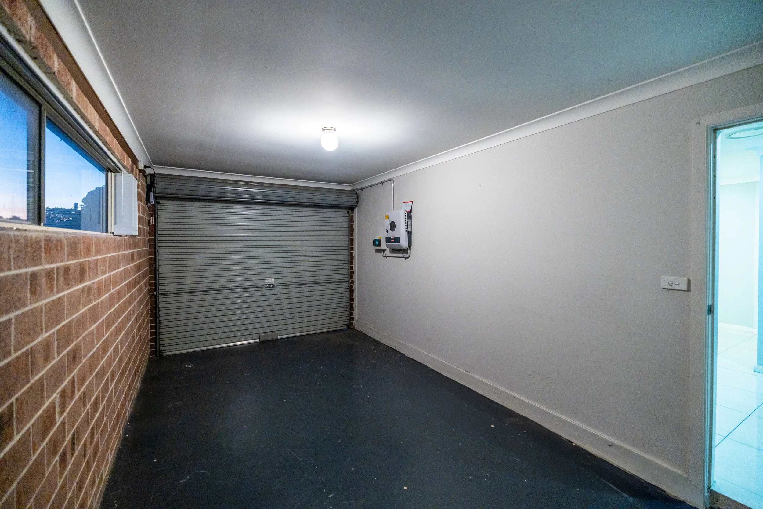Empty garage with a brick wall on the left, a white wall with electrical equipment on the right, a closed garage door at the back, and a small window on the left.