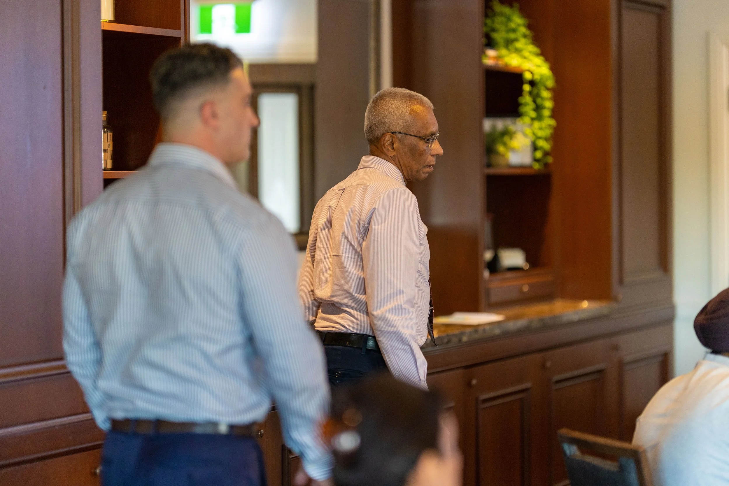 Two men standing indoors, one elderly with gray hair and glasses, the other younger with short dark hair, both dressed in collared shirts, in a room with wood-paneled walls and a granite countertop.
