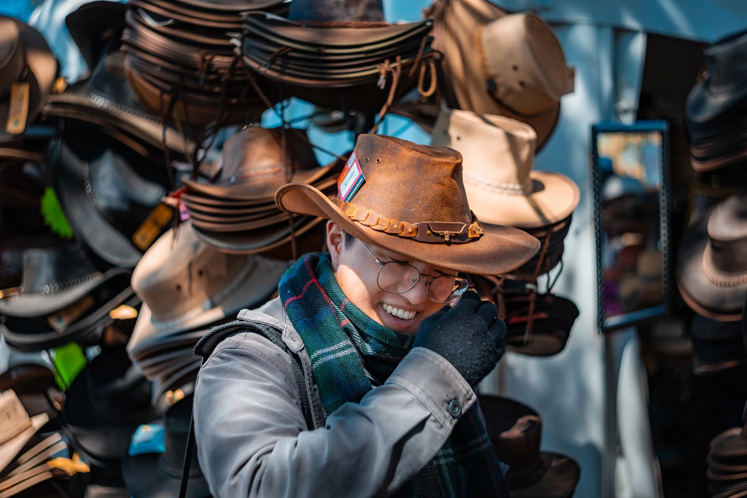 A person wearing glasses, a plaid scarf, and a gray coat trying on a brown cowboy hat in front of a display of various hats at an outdoor market.