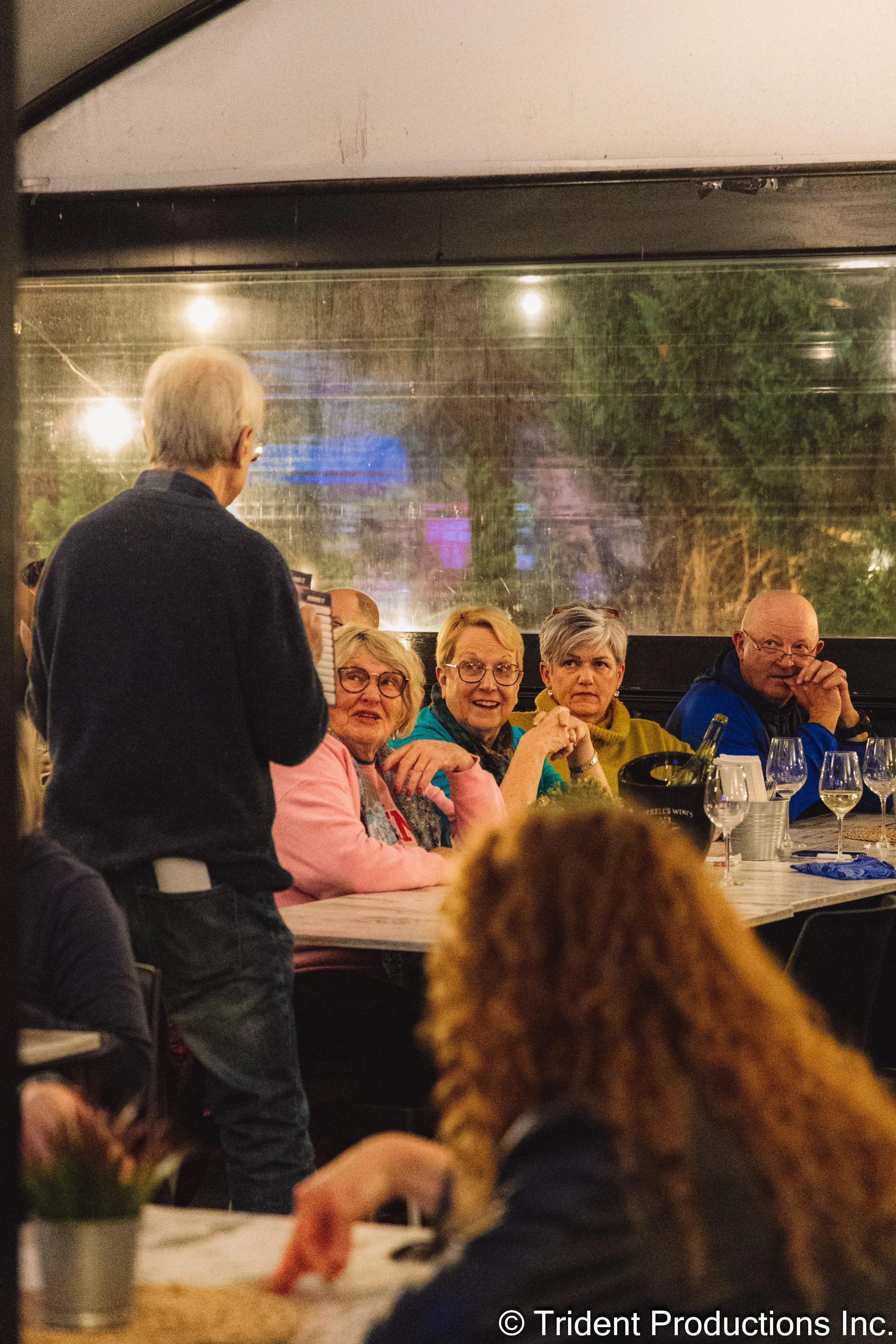 A group of four women sitting at a dinner table in a restaurant, listening to a person standing and speaking to them, with a window behind them showing colorful reflections outside.