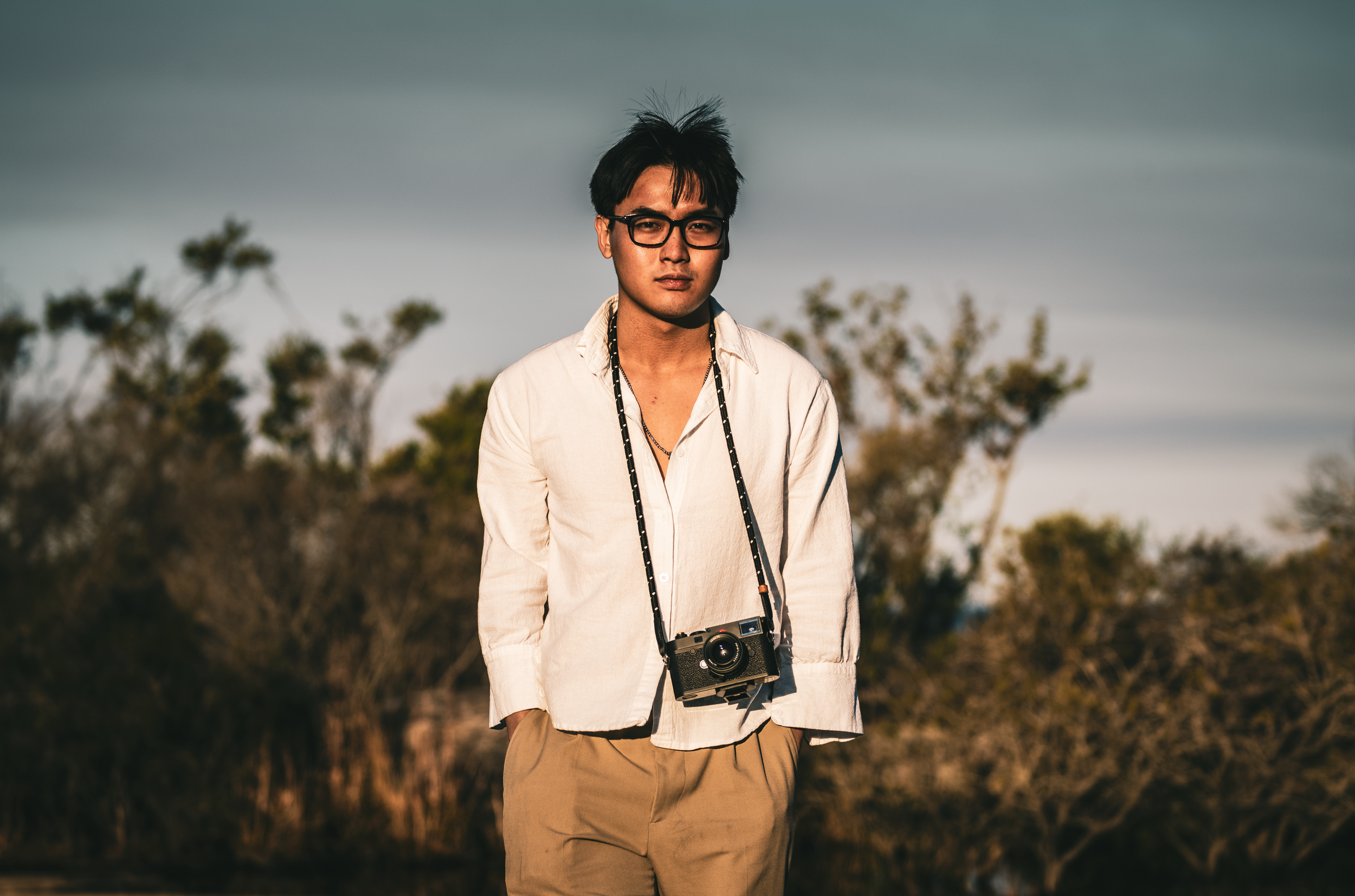 Young man with black hair, glasses, white shirt, beige pants, and a camera around his neck standing outdoors with trees in the background during sunset.