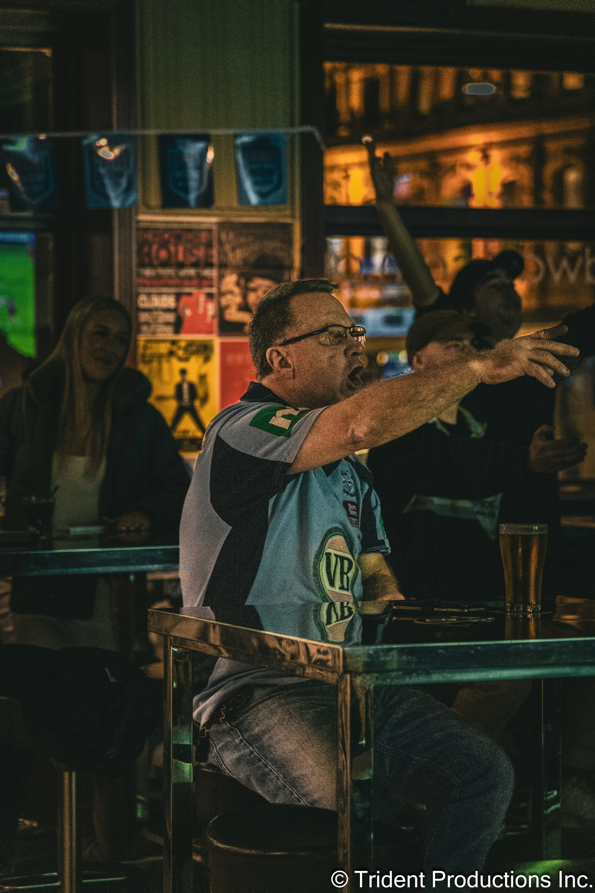 A man in a sports jersey shouting and gesturing with his hand in a bar or pub with a dimly lit background, other people sitting and watching, posters on the wall, and theatrical lighting.