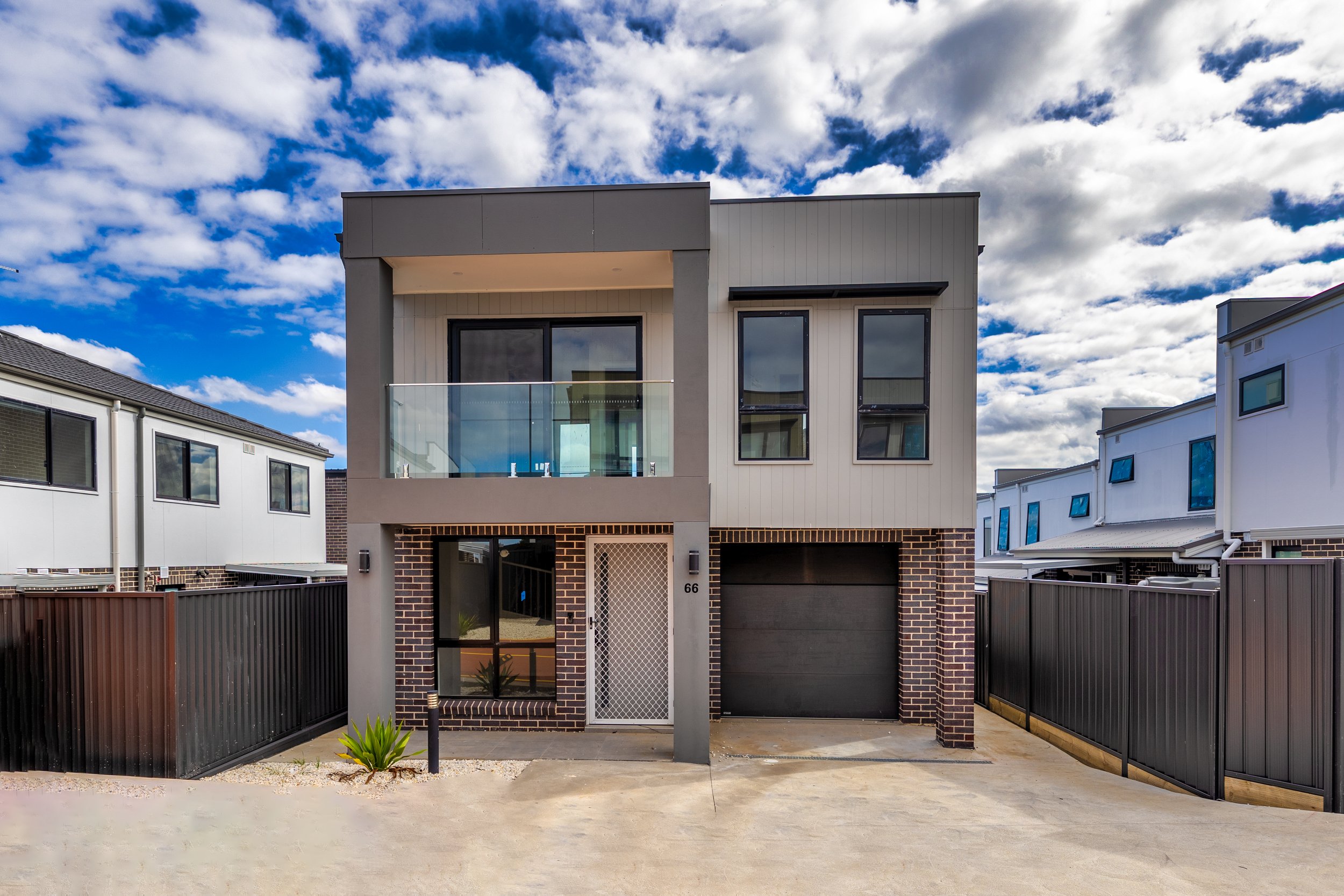 Modern two-story house with brick and white panel exterior, black garage door, and glass balcony in front. The house number is 66, and there is a small plant by the entrance. Fenced yard with neighboring houses on both sides under a partly cloudy sky