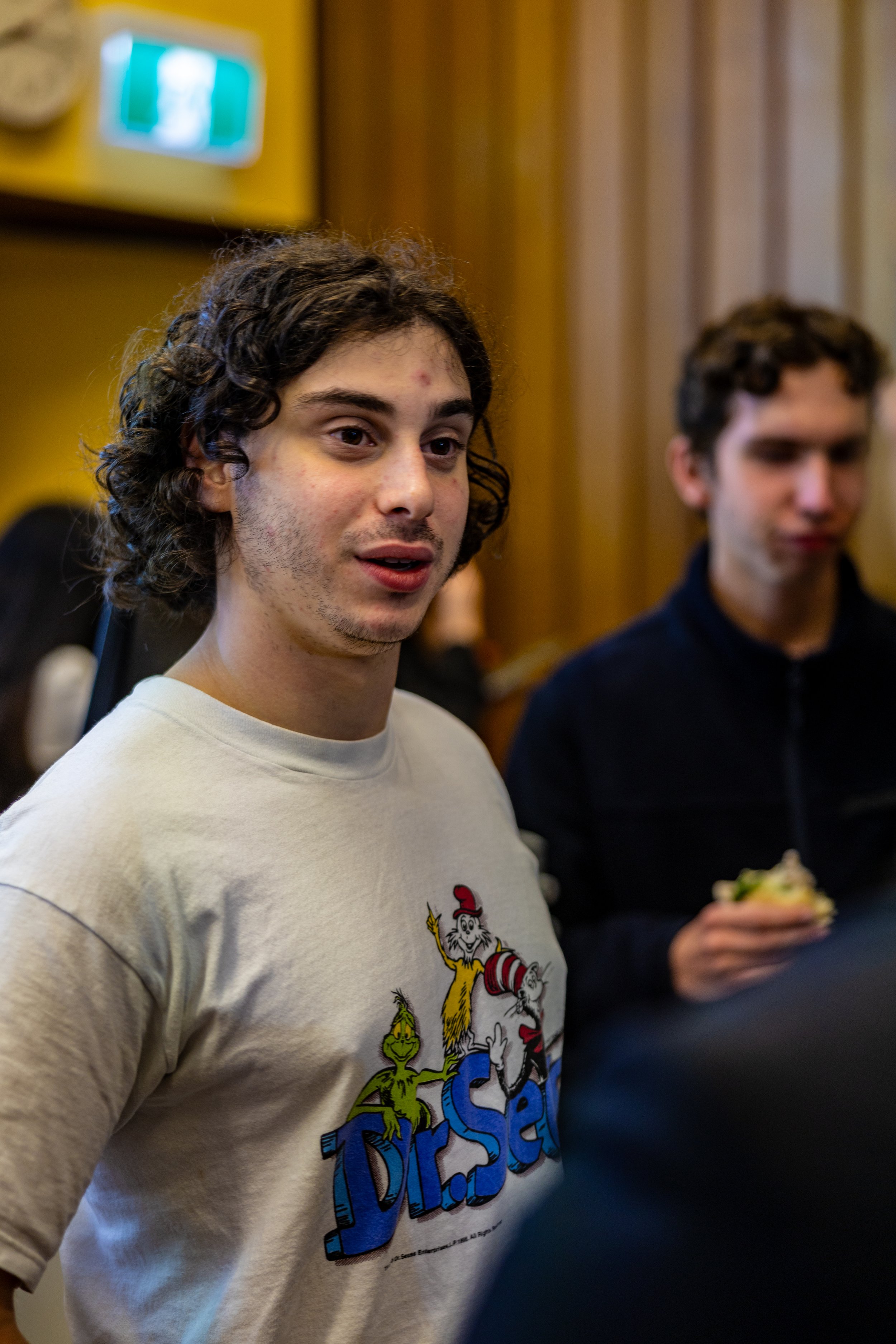 A young man with curly brown hair and light facial hair wearing a Dr. Seuss T-shirt, standing indoors with wooden paneling and other people in the background.