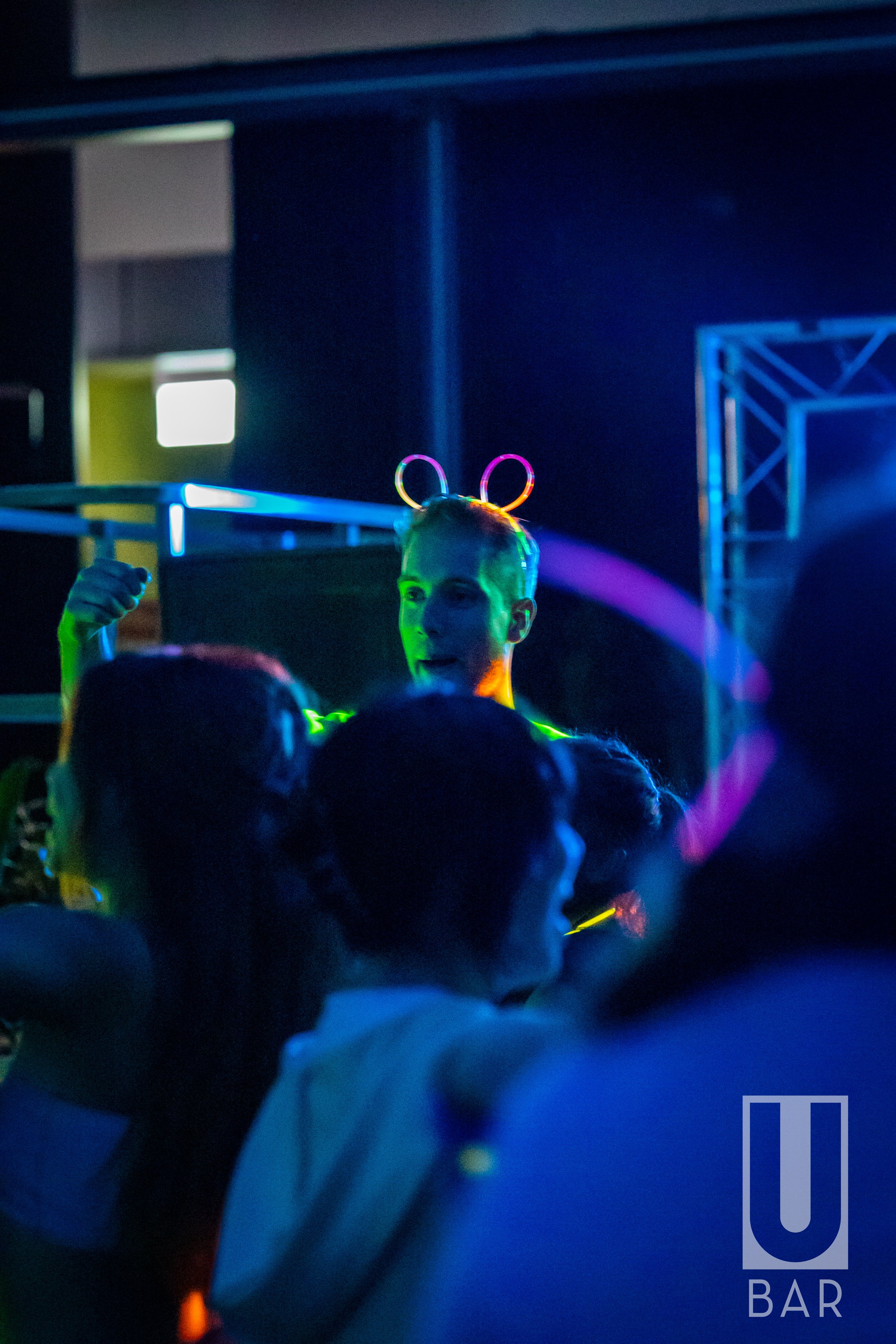 Person with glowing bunny ears headband at a party or club, surrounded by young people, colorful lights, and a dark background.