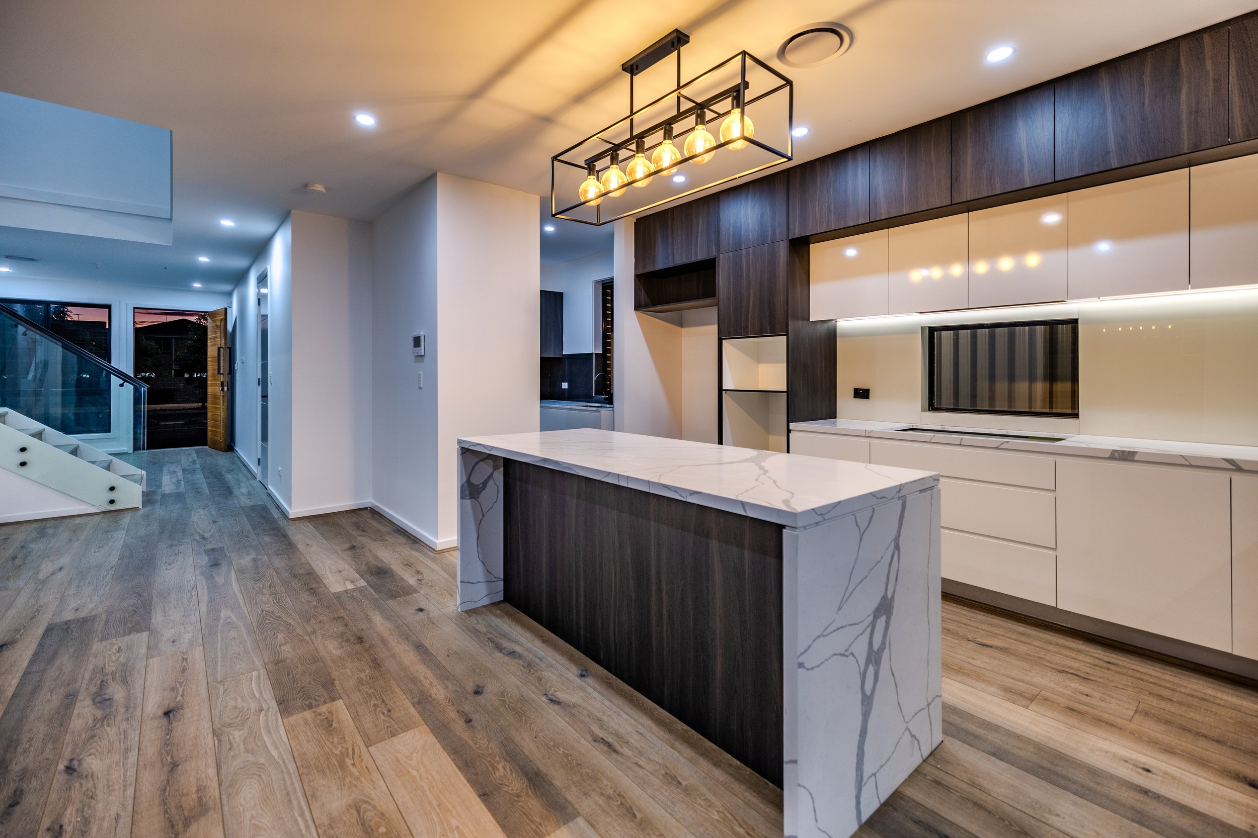 Modern kitchen with a white marble island, dark wood cabinets, white upper cabinets, and a black lighting fixture with exposed bulbs, with wooden flooring and a view of a living area and staircase.
