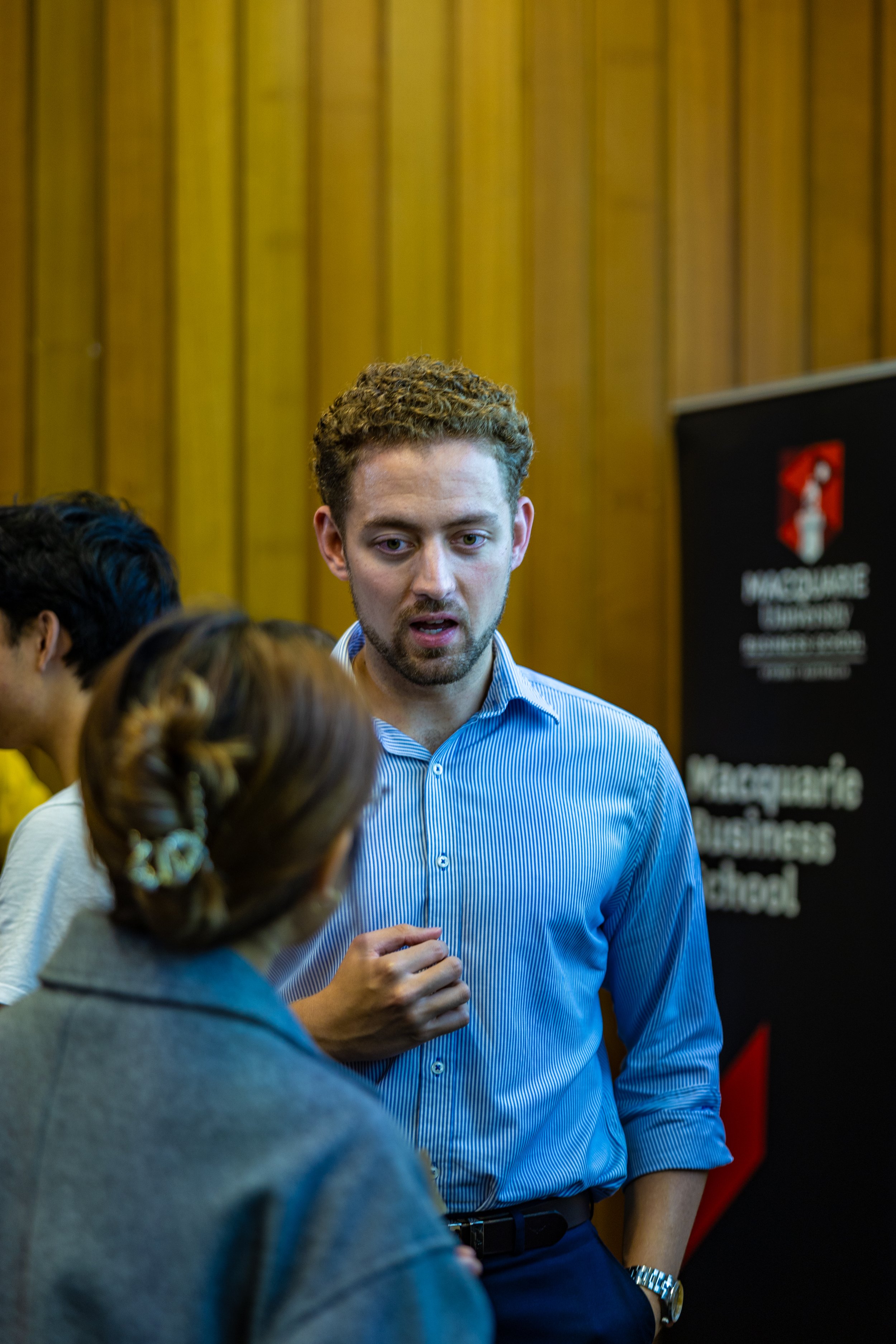 A young man with curly hair, wearing a blue striped shirt, speaking to a woman with braided hair in a grey jacket at an event with a black and red banner reading 'Macquarie Business School' in the background.