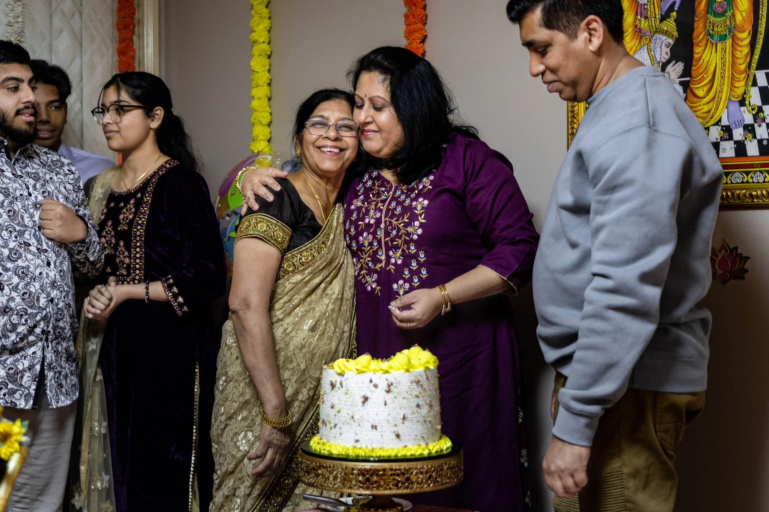 Group of people celebrating a birthday, with a woman in a gold saree hugging another woman in a purple kurta, standing near a cake with yellow flowers.