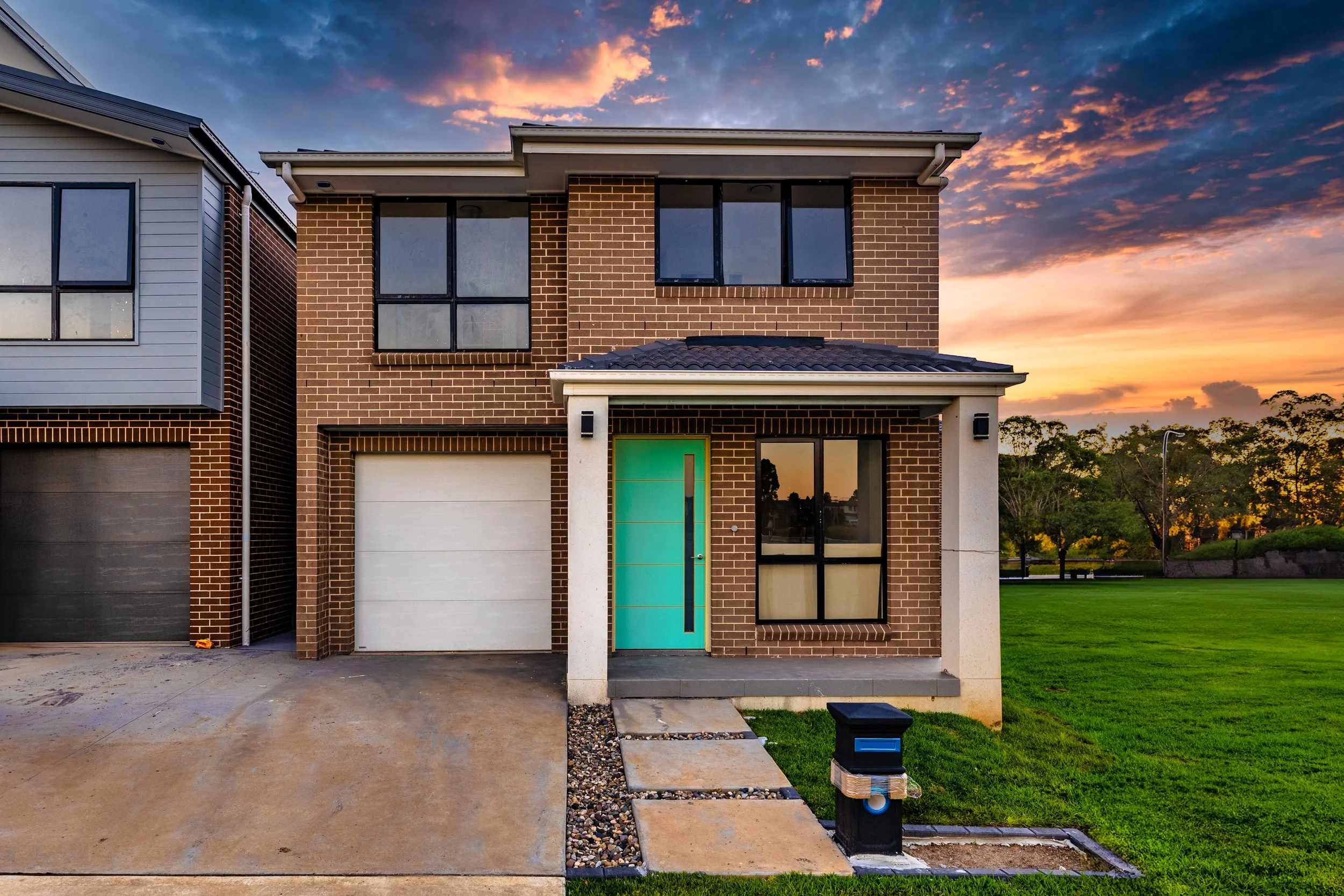 Modern two-story brick house with a green front door at sunset. The house has large windows, a white garage door, a concrete driveway, and a small front porch with a pathway leading to the door. There is a mailbox at the sidewalk and a green lawn wit
