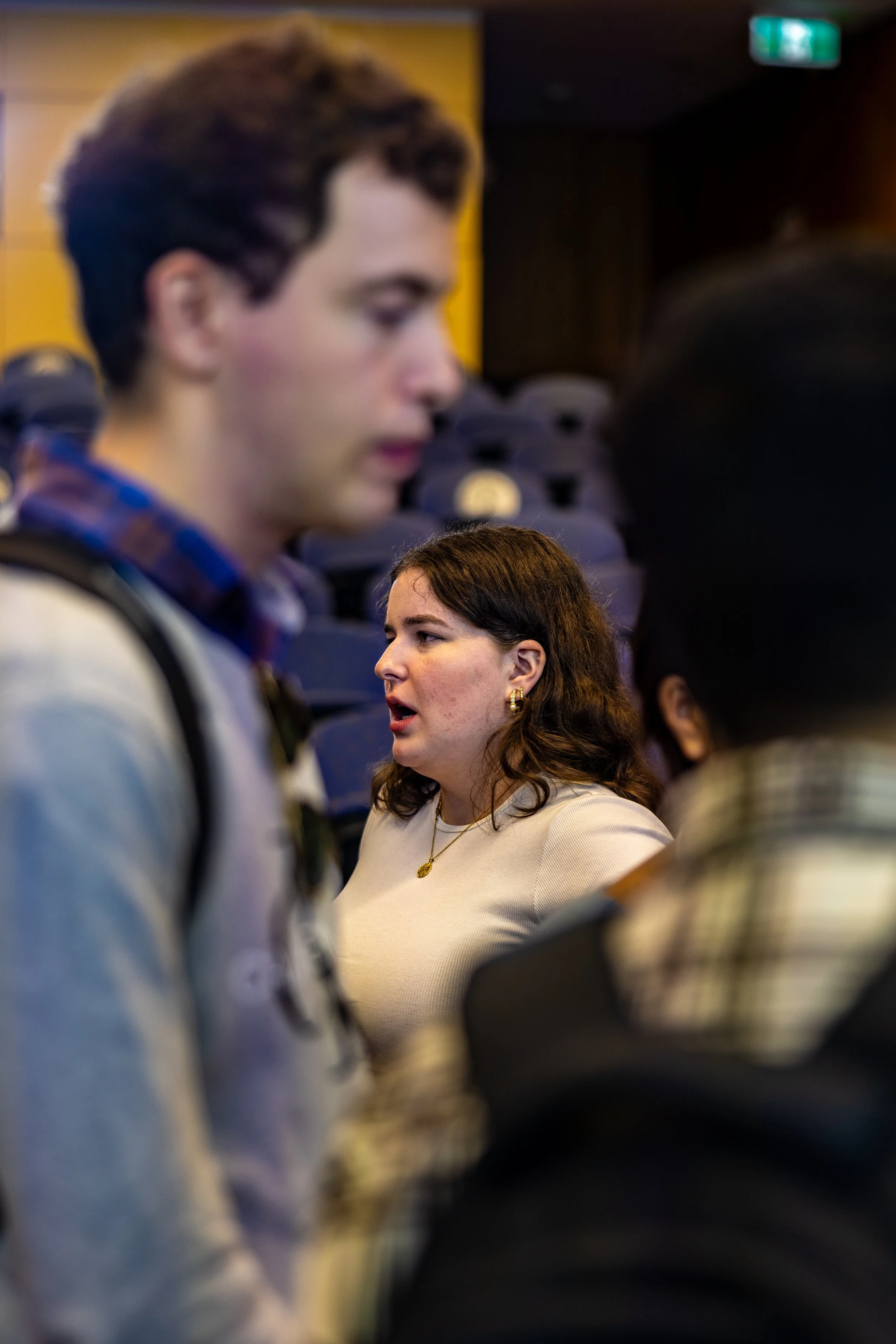 A woman with dark brown hair and earrings appears to be speaking or reacting, with a young man with short brown hair and a backpack in the foreground, and rows of empty blue chairs in the background.