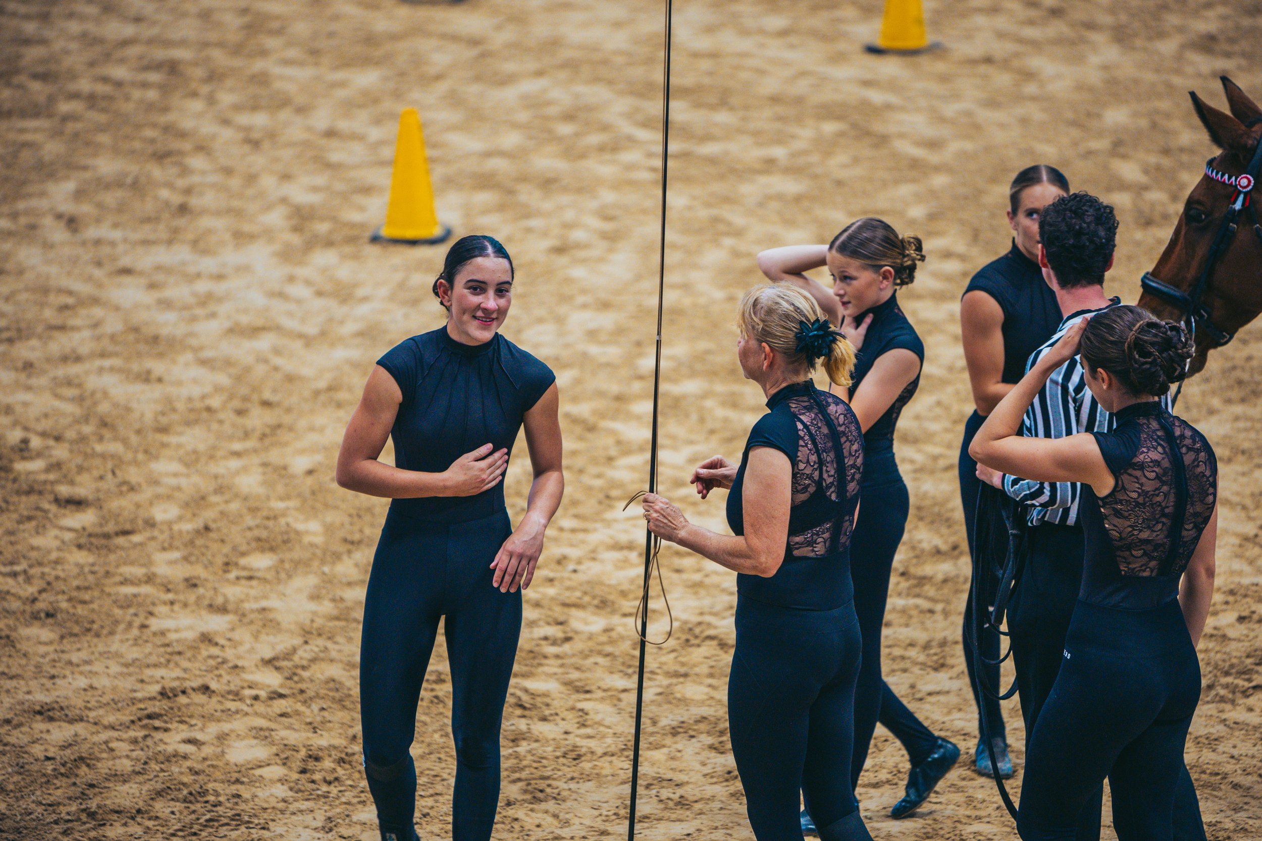 A group of young women in black outfits standing and talking in an indoor riding arena with a horse and yellow cones in the background.