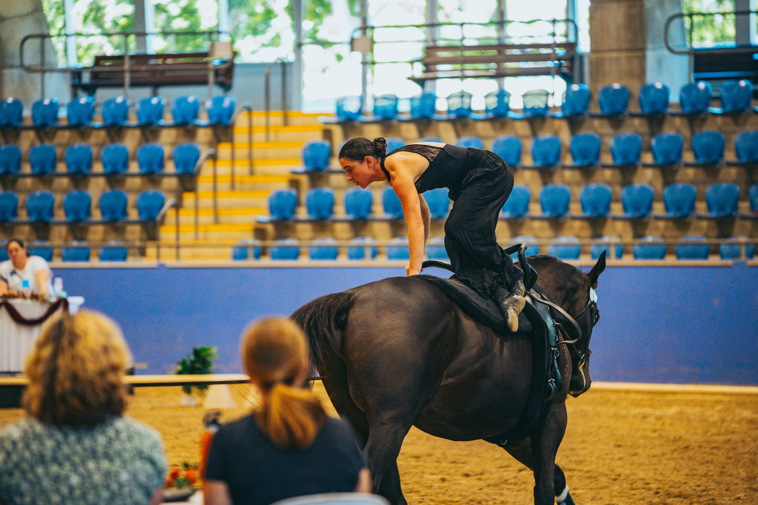 A woman in black attire balancing on a horse in an indoor arena, with spectators watching.