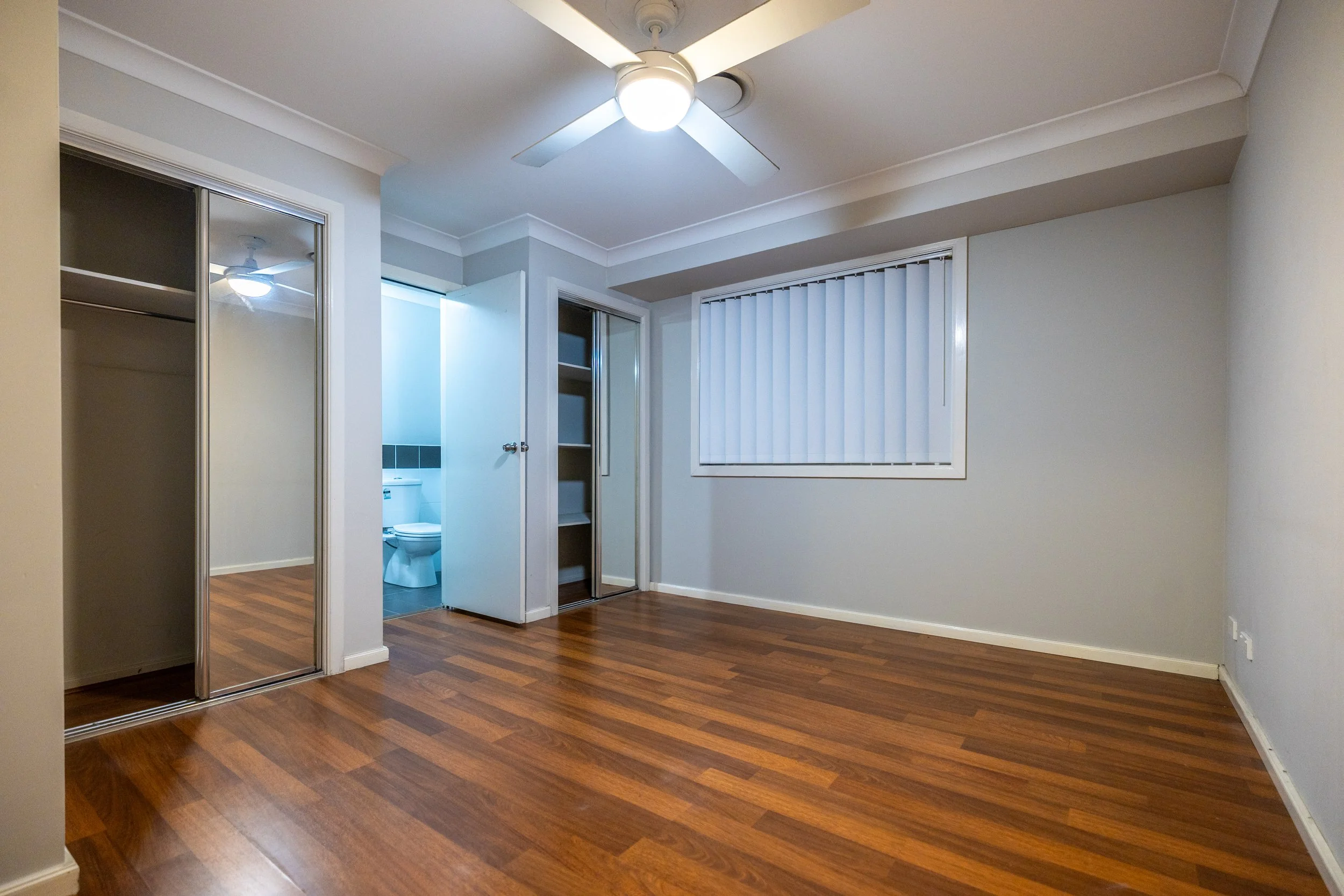 Empty room with wood flooring, a ceiling fan, a window with vertical blinds, a closet with mirrored sliding doors, and a doorway leading to a bathroom with a toilet.
