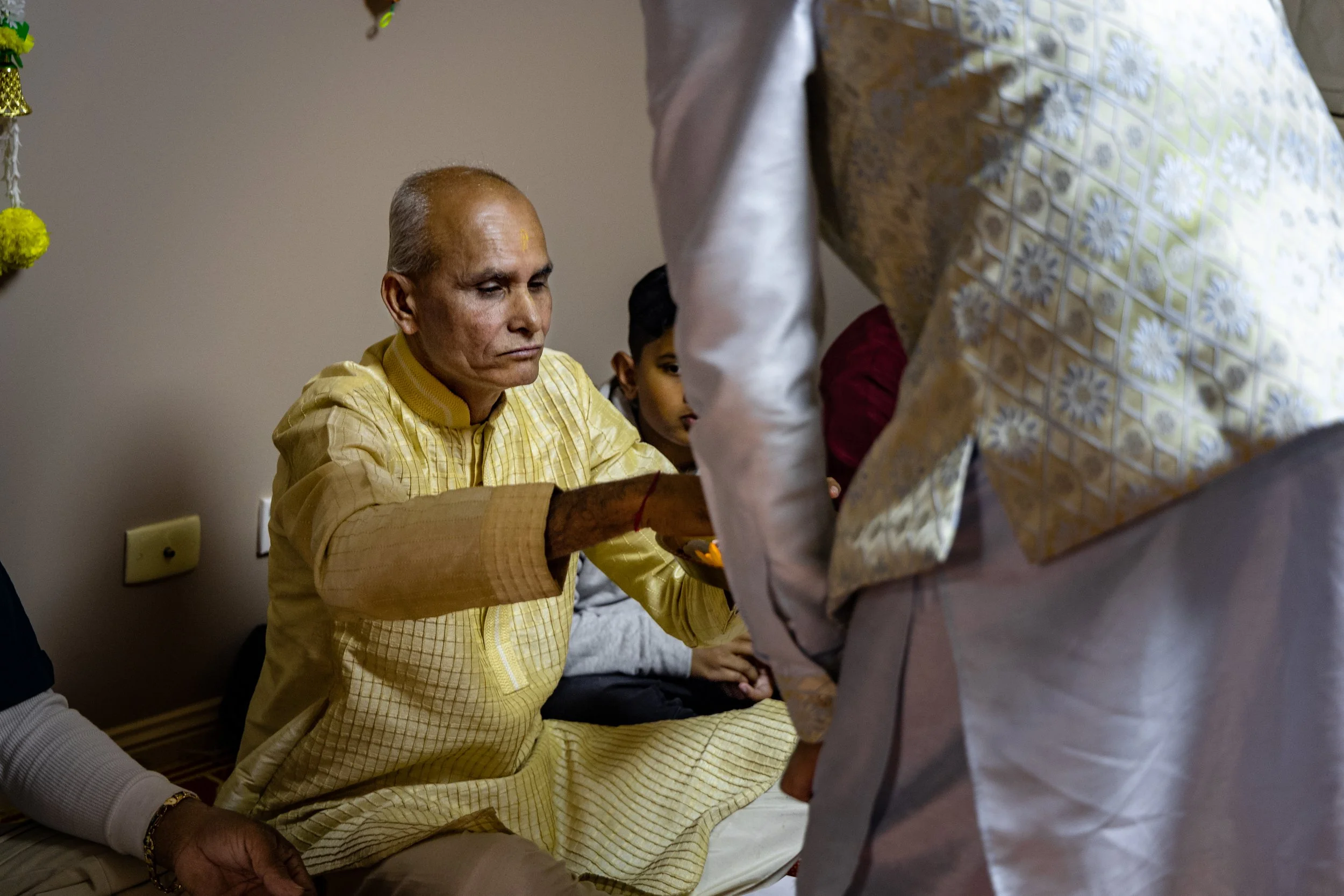 An elderly man dressed in yellow traditional Indian attire performing a religious ritual, with other people sitting nearby, one in a gray sweater and the other a young boy, participating in the ceremony.