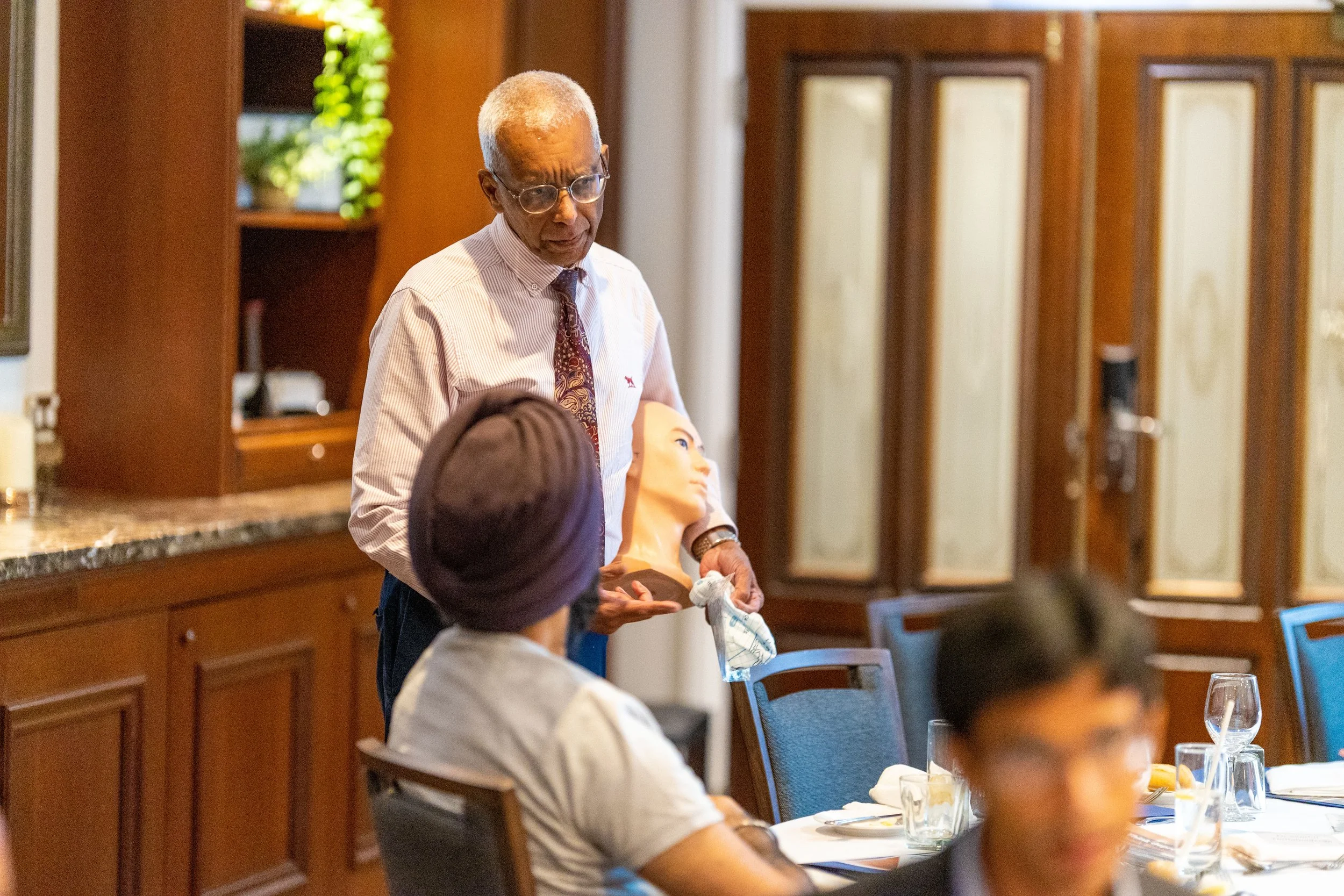 An elderly man with gray hair and glasses is holding a medical mannequin head and speaking to a group of people seated at a dining table in a room with wooden cabinets.