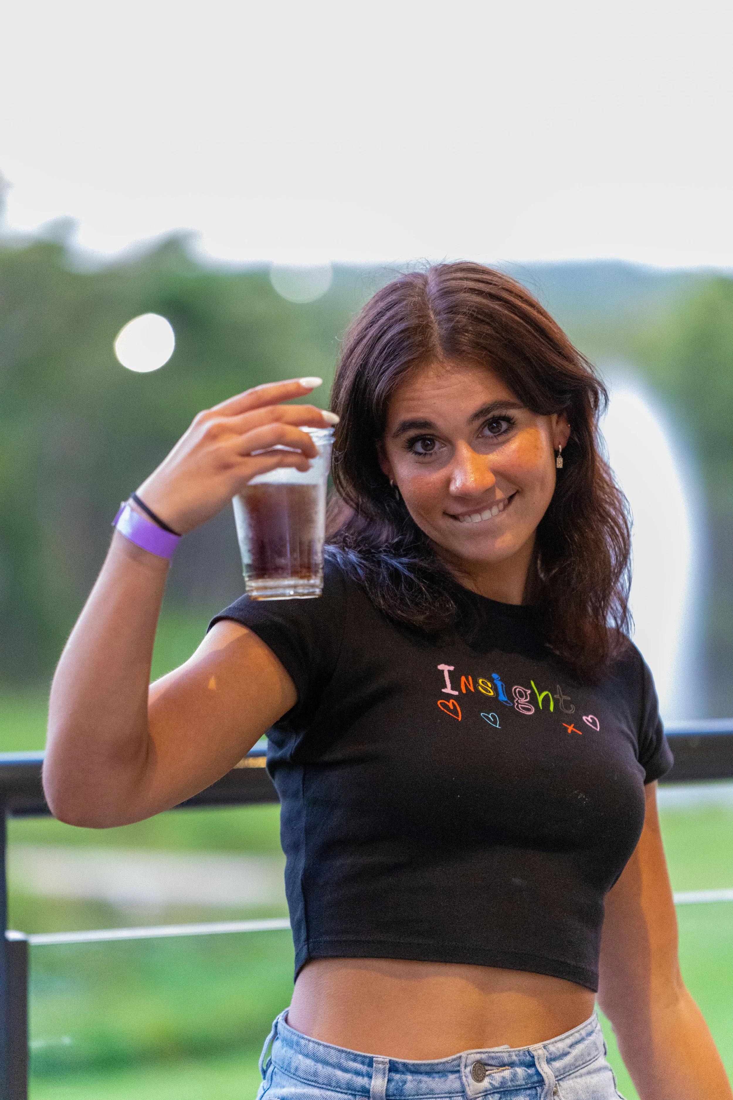 Young woman with dark hair holding a glass of dark beverage outside with green trees and a water fountain in the background.
