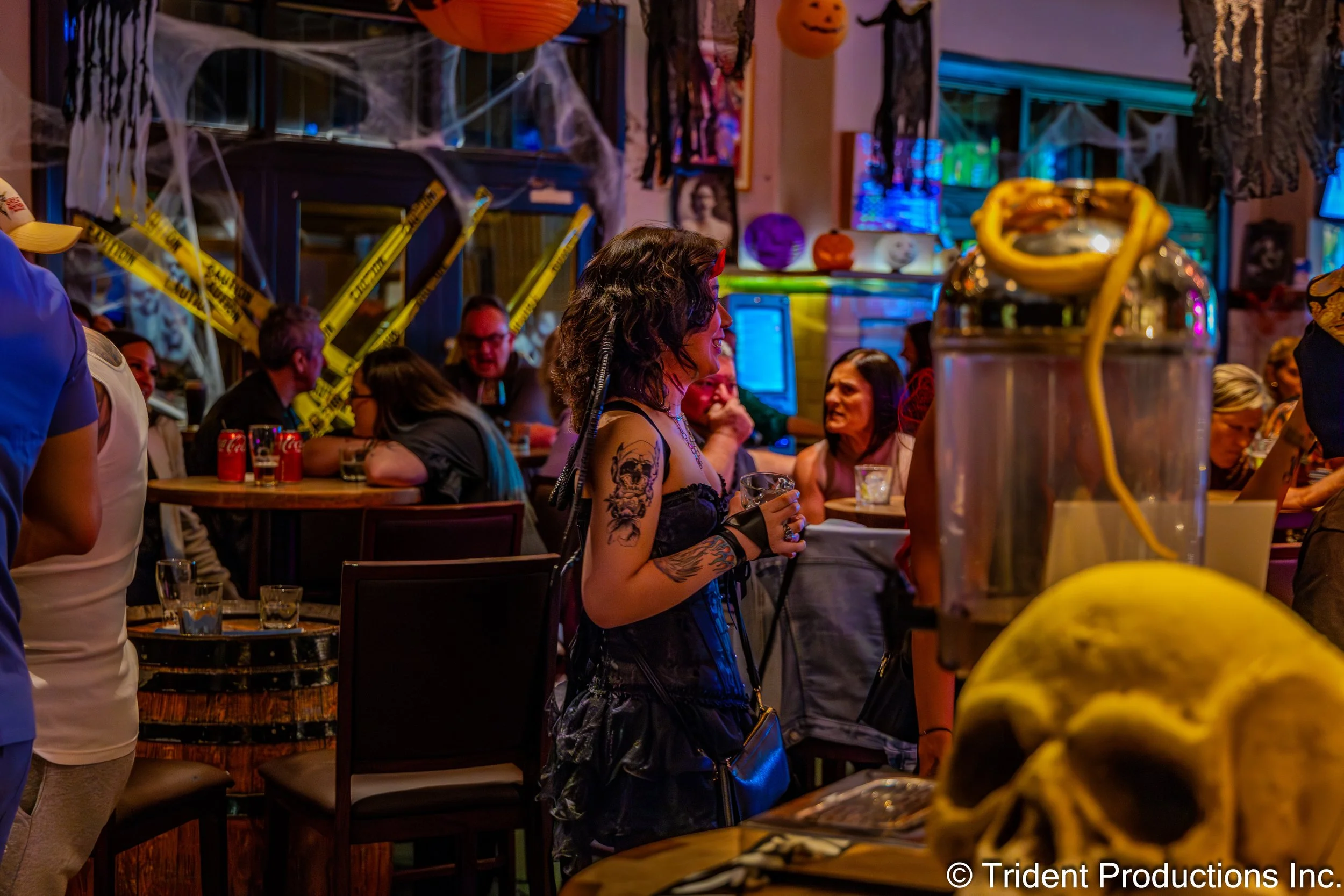A Halloween party scene with people socializing. There are spooky decorations like cobwebs, 'Caution' tape, and pumpkins. A woman with curly hair and tattoos is in the center, holding a drink. In the foreground, there's a large transparent container 