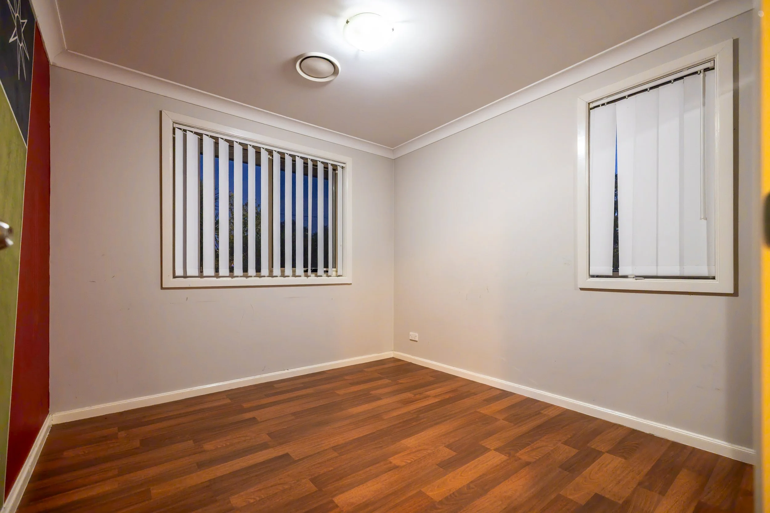 Empty room with white walls, hardwood floor, two windows with vertical blinds, and ceiling light fixture.