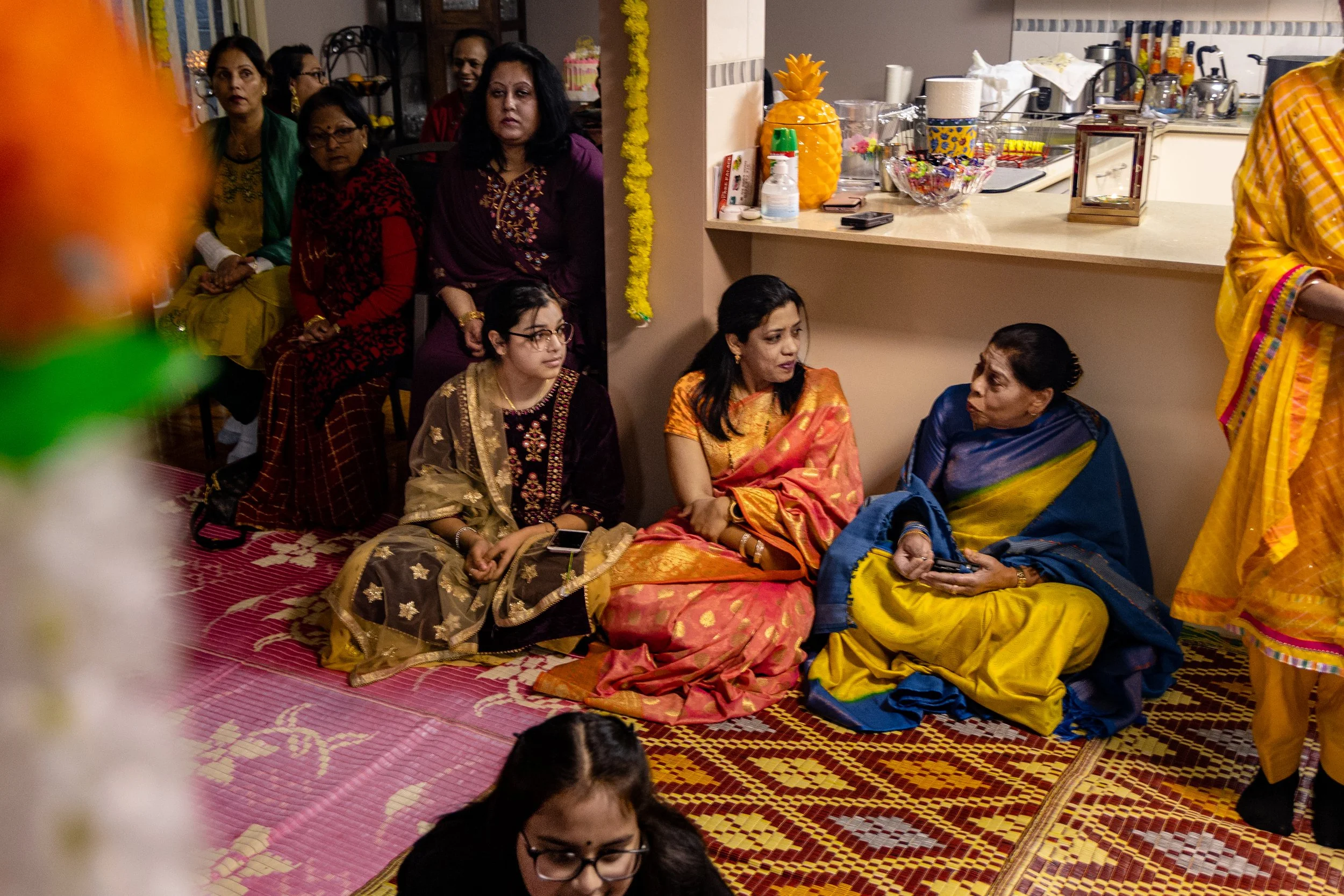 Group of women at an indoor Indian celebration, some sitting and others standing, wearing colorful traditional sarees and kurta, on a patterned carpet, with a kitchen counter visible in the background.