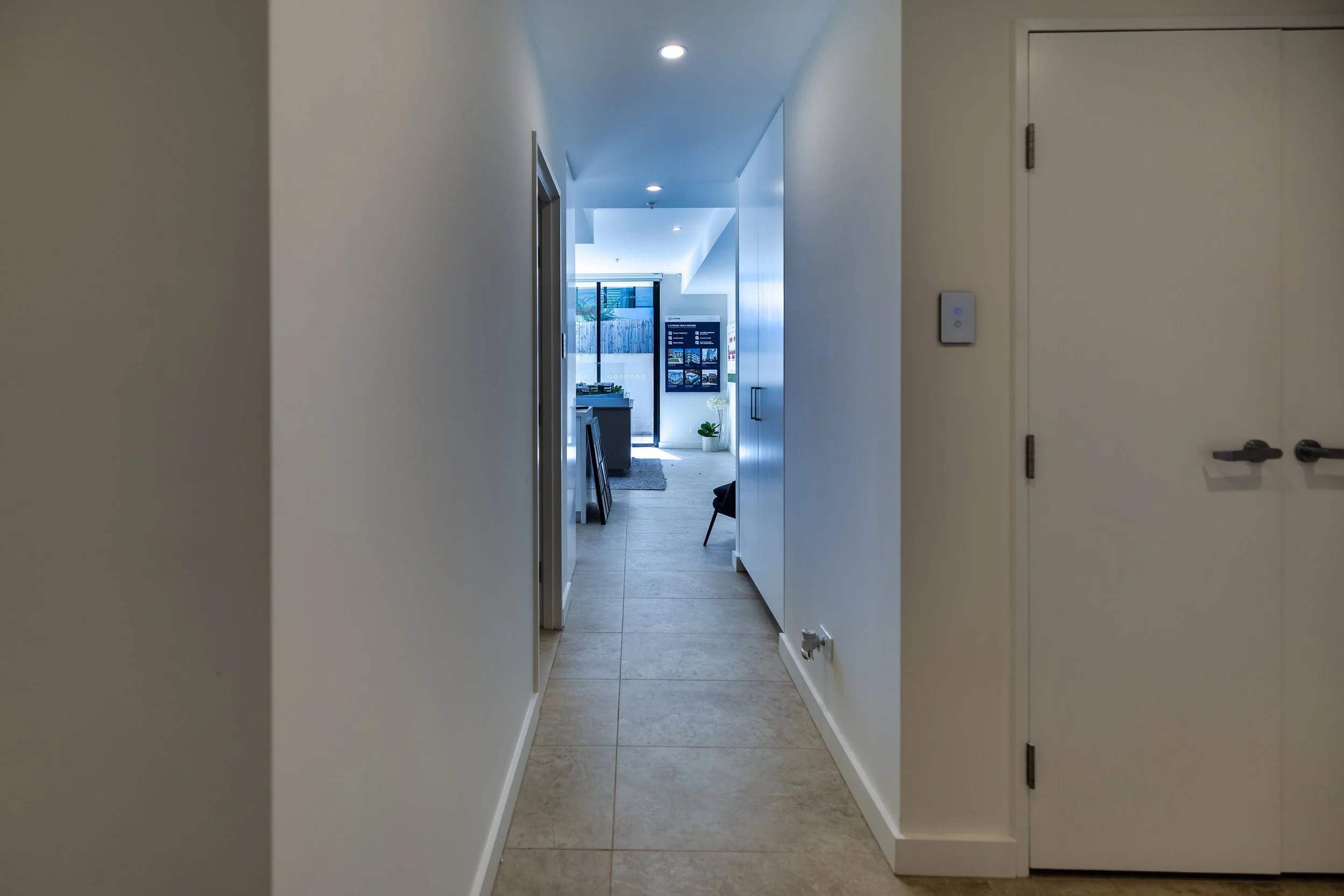 View of a modern apartment hallway leading to a living room area with sliding glass doors, a television, a plant, and some furniture.