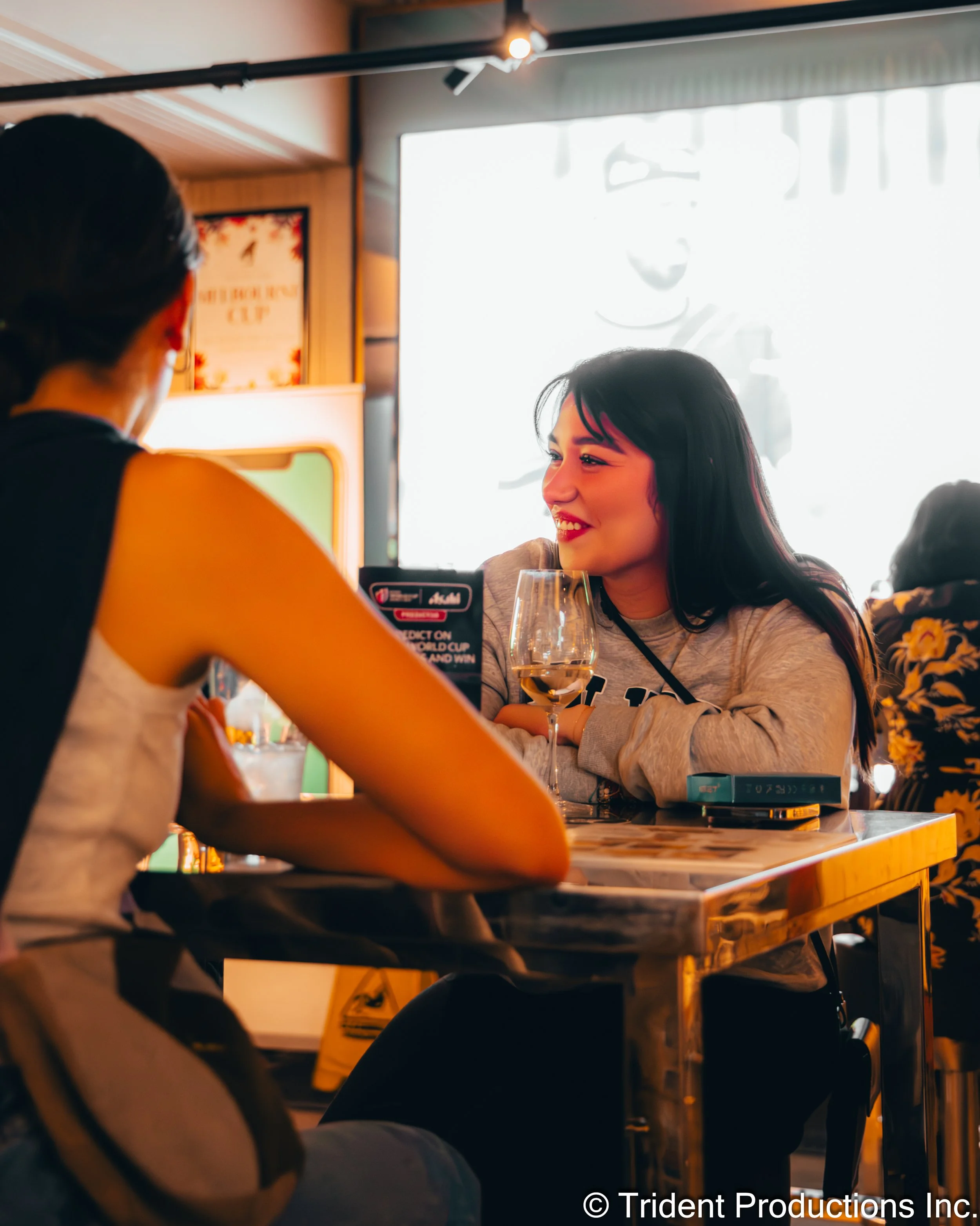 Two women sitting at a table, talking, with one smiling and holding a glass of white wine, in a restaurant or cafe with a bright window in the background.