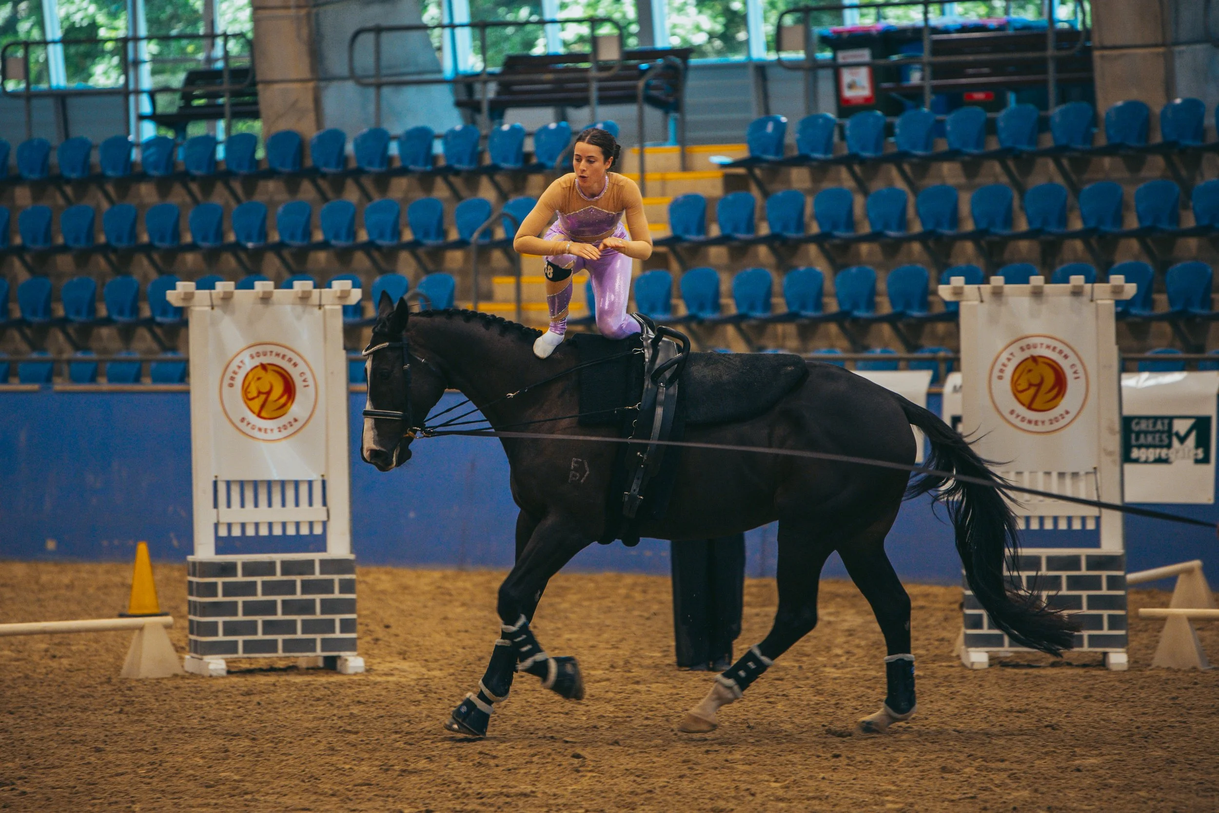 A woman dressed as a princess balancing on a black horse in an indoor arena, with blue empty seats in the background, during a horse riding event.