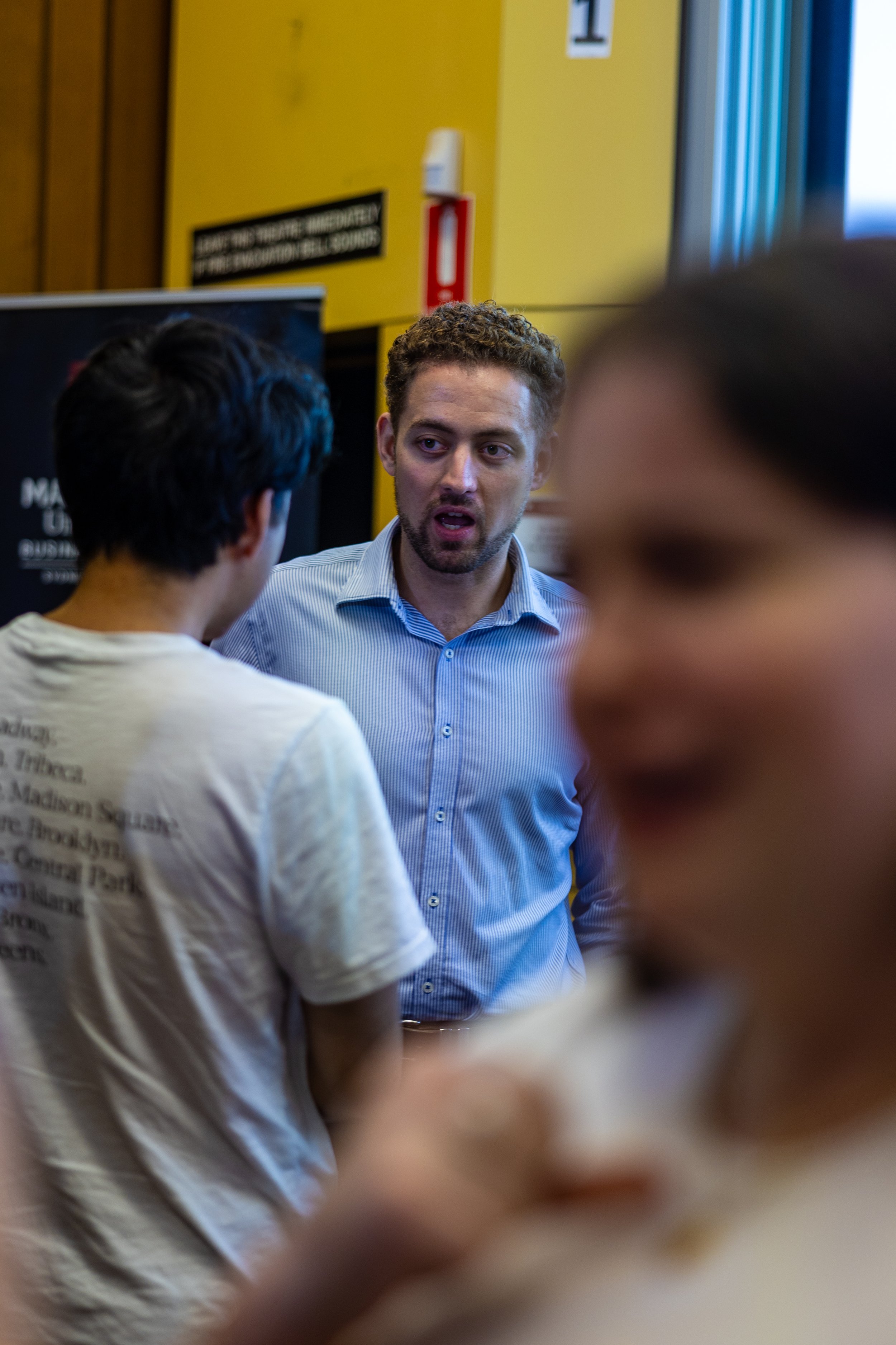 A man with curly hair and a beard, wearing a blue striped shirt, appears to be speaking to two other people in a crowded room.