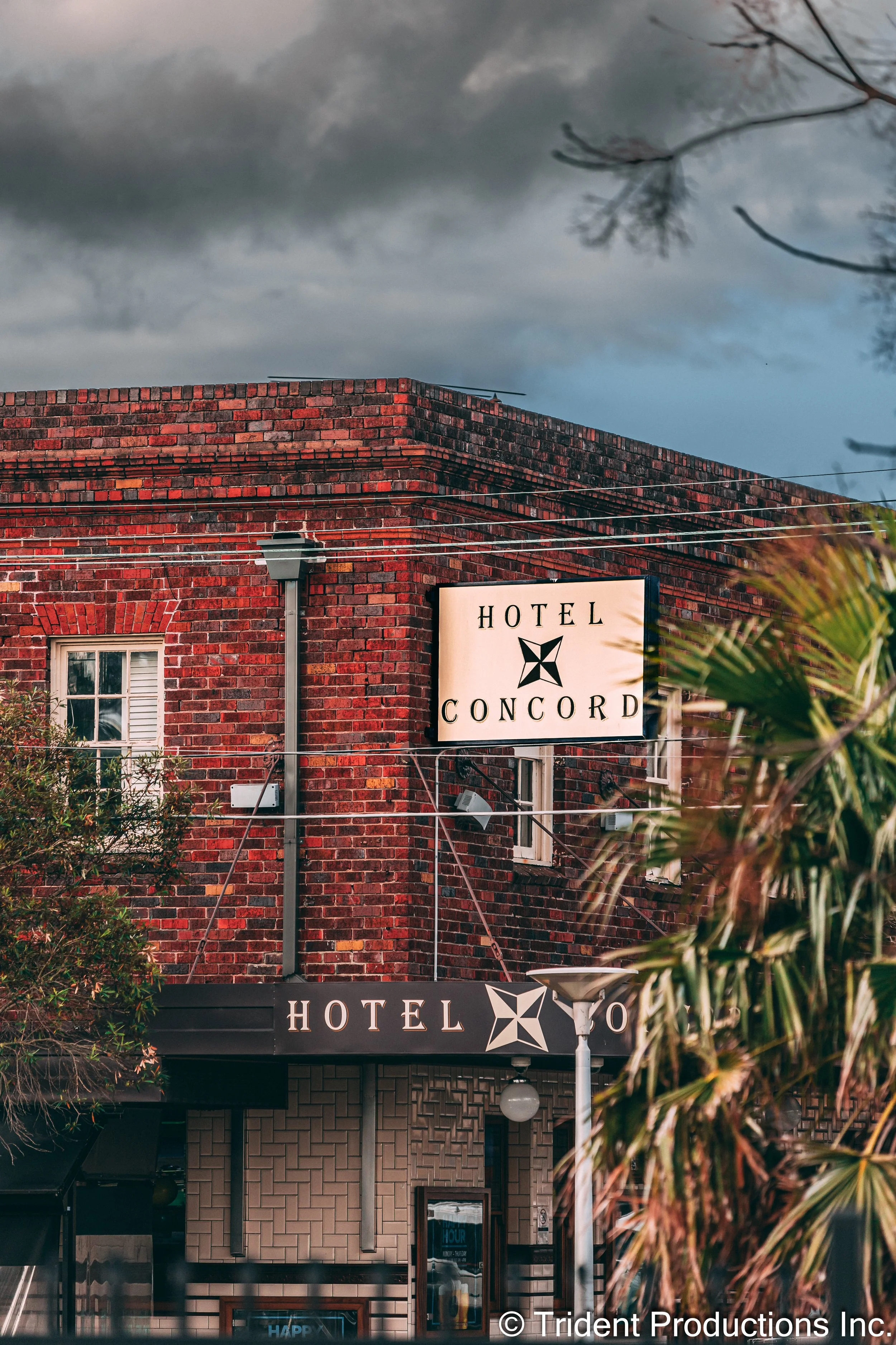 Red brick hotel building with a sign reading 'Hotel Concord' and a banner with the same name and logo, with cloudy sky and some trees in the foreground.