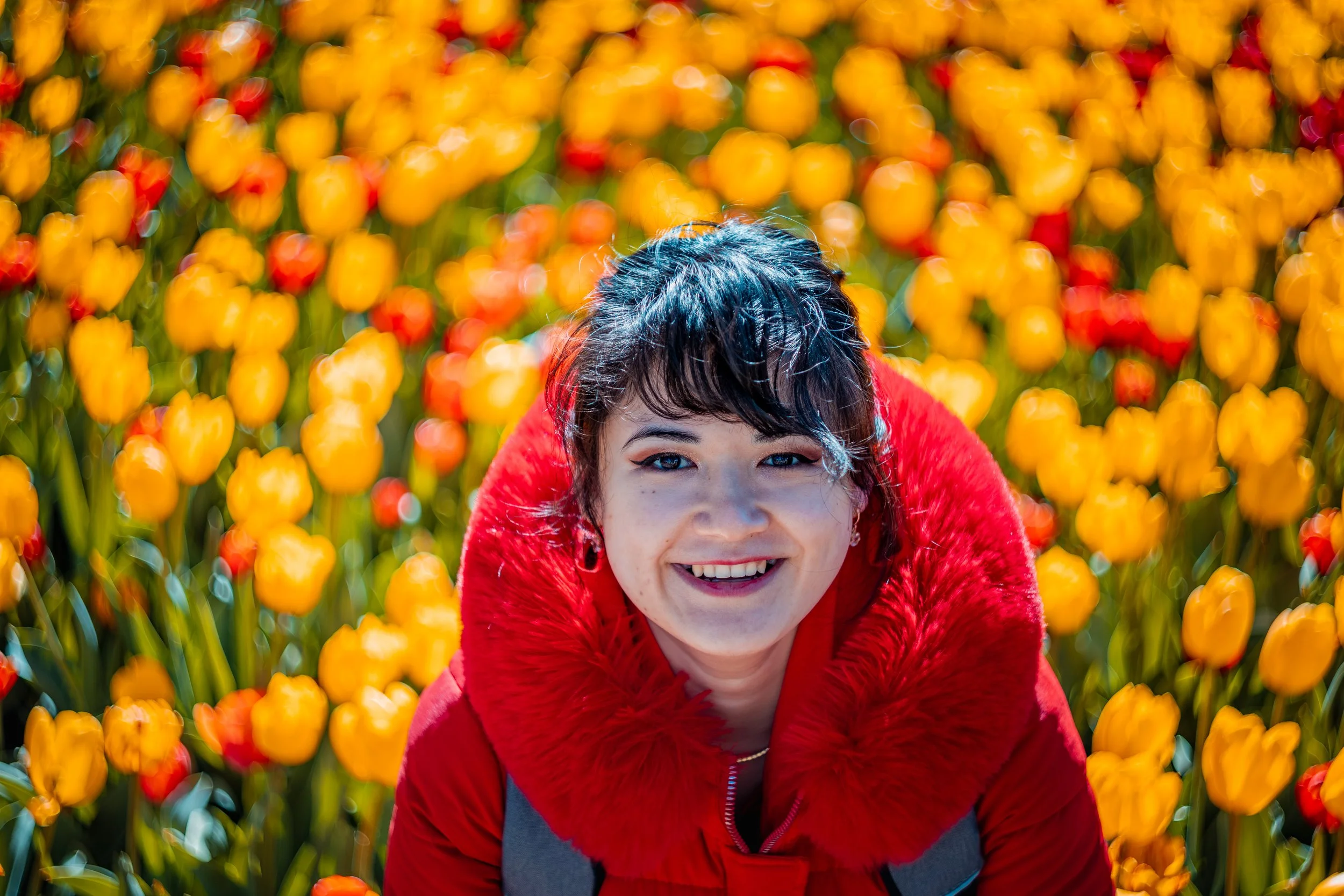 A smiling woman wearing a red coat with a fur collar in a field of yellow and red tulips.