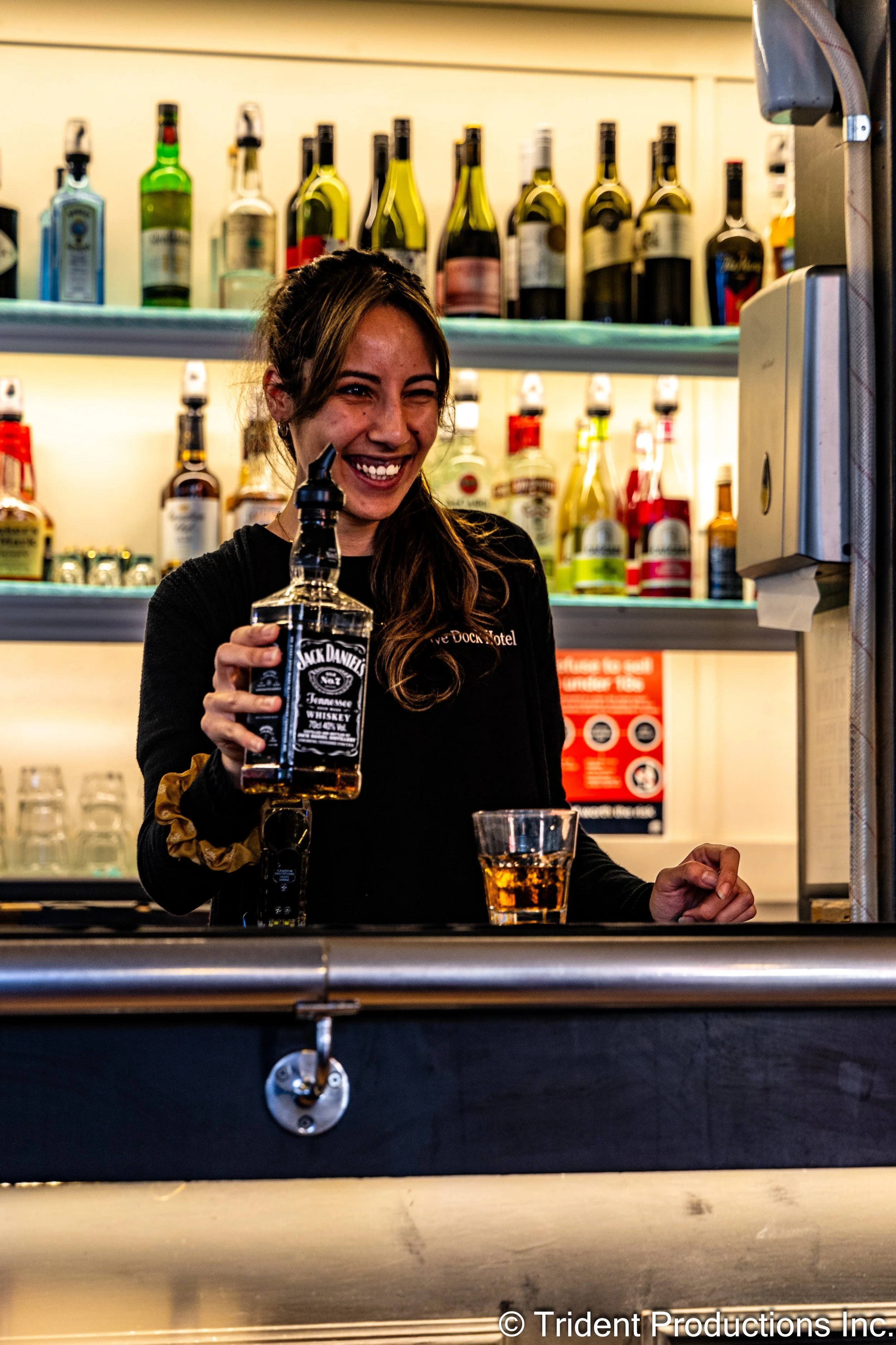 A woman smiling and holding a bottle of Jack Daniel's whiskey behind a bar counter, with liquor bottles on shelves in the background.