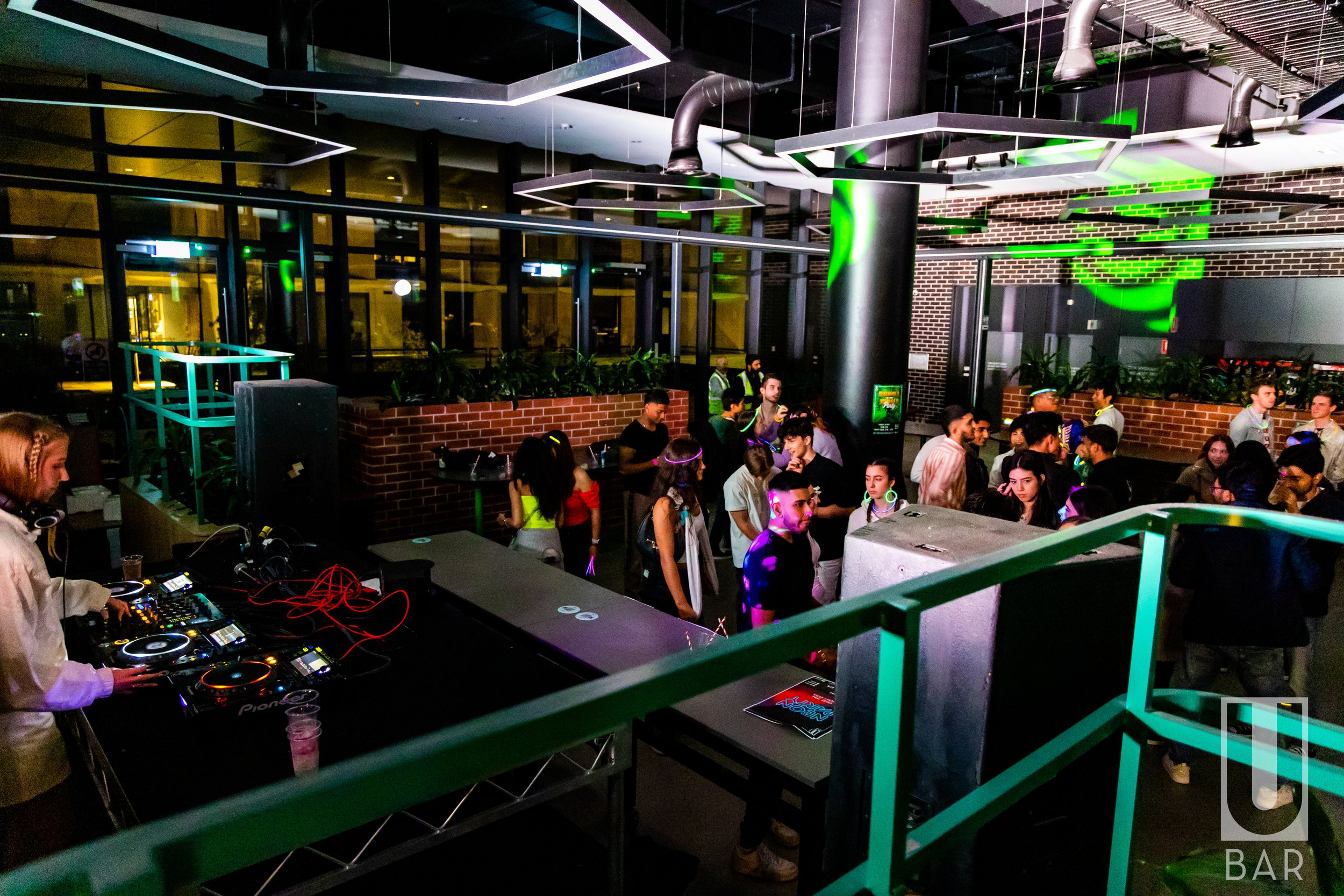 A crowded nightclub with people dancing and socializing, a DJ performing at a booth, brick walls, green lighting, and neon accents.