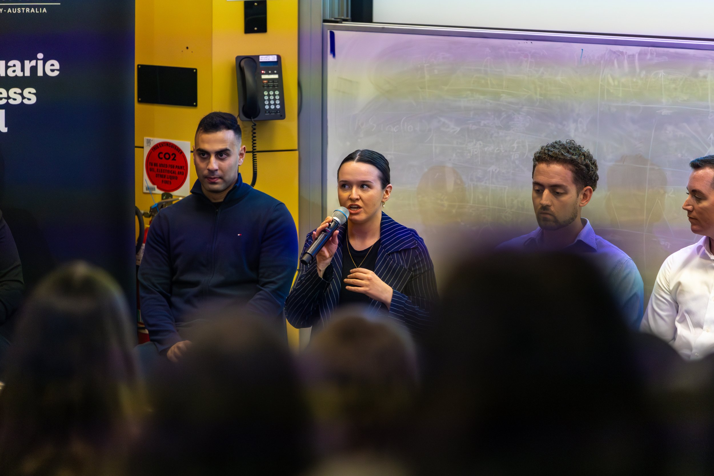 A woman speaking into a microphone at a panel discussion or presentation, with three men sitting beside her in a classroom or conference room setting.