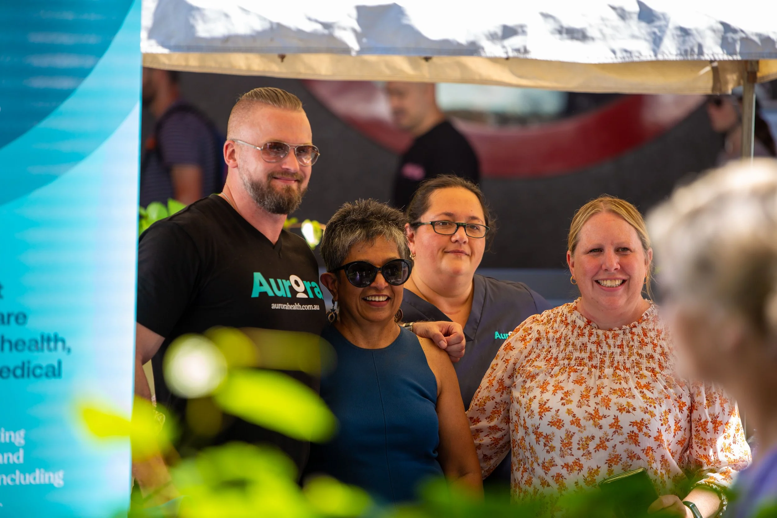 Four people smiling at a community event or health fair, standing behind a booth with a blue banner.