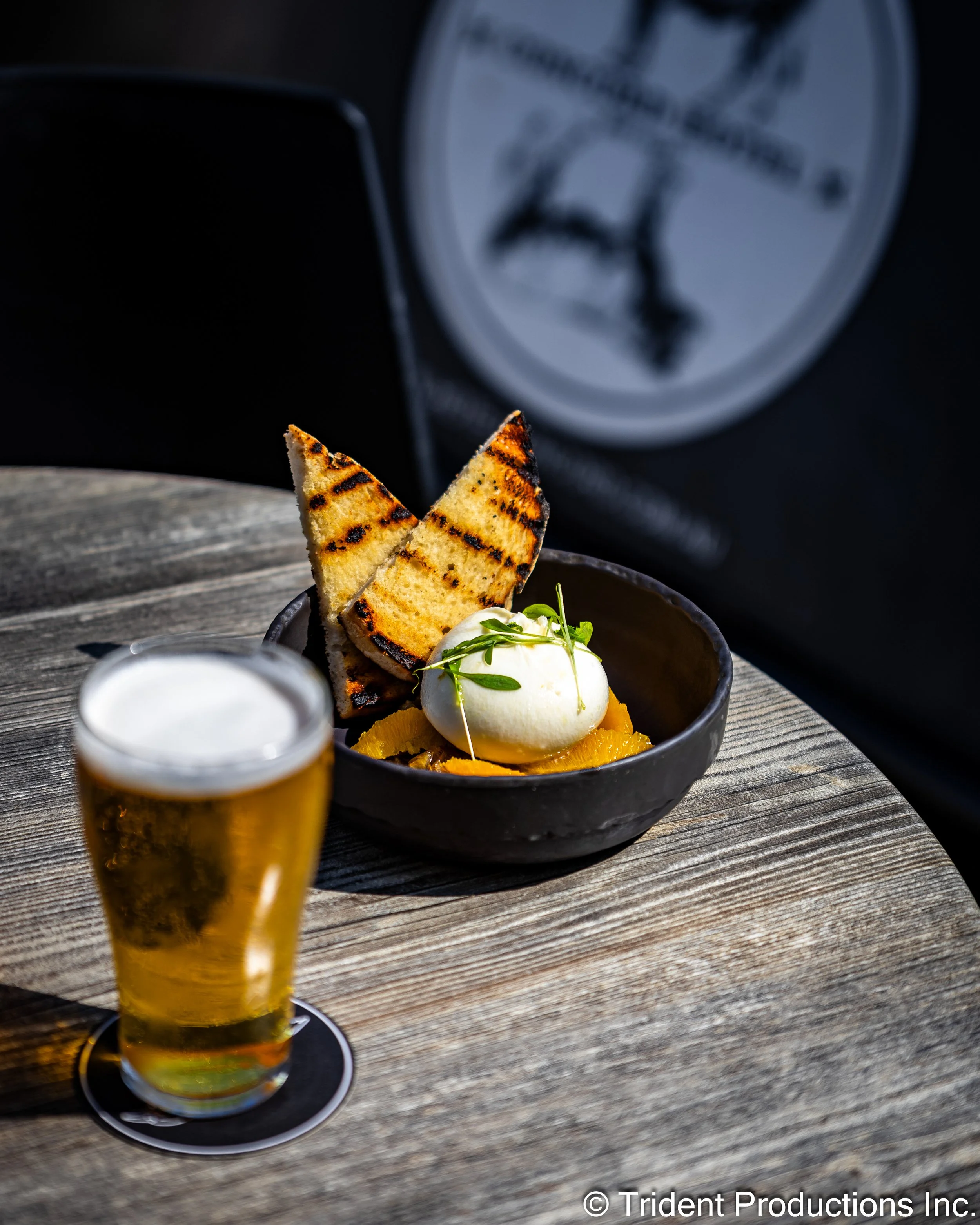 A bowl of dessert with grilled toast, a scoop of ice cream, and orange slices, with a glass of beer on a wooden table.