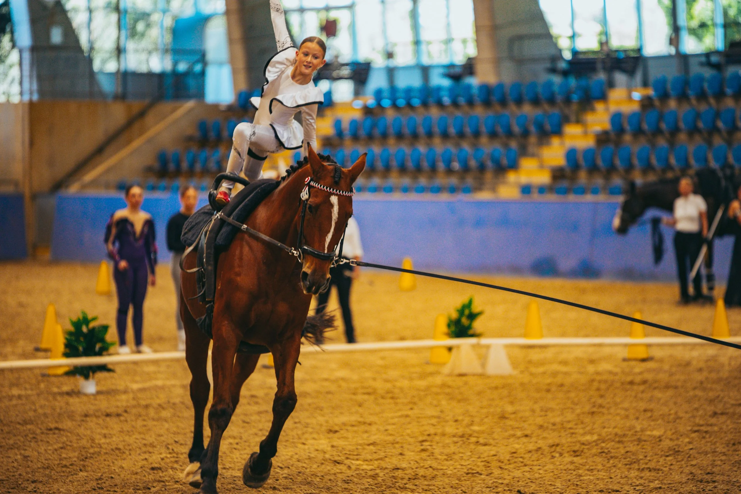 A young girl in an equestrian outfit performing a trick on a brown horse inside an indoor riding arena, with spectators and officials in the background.