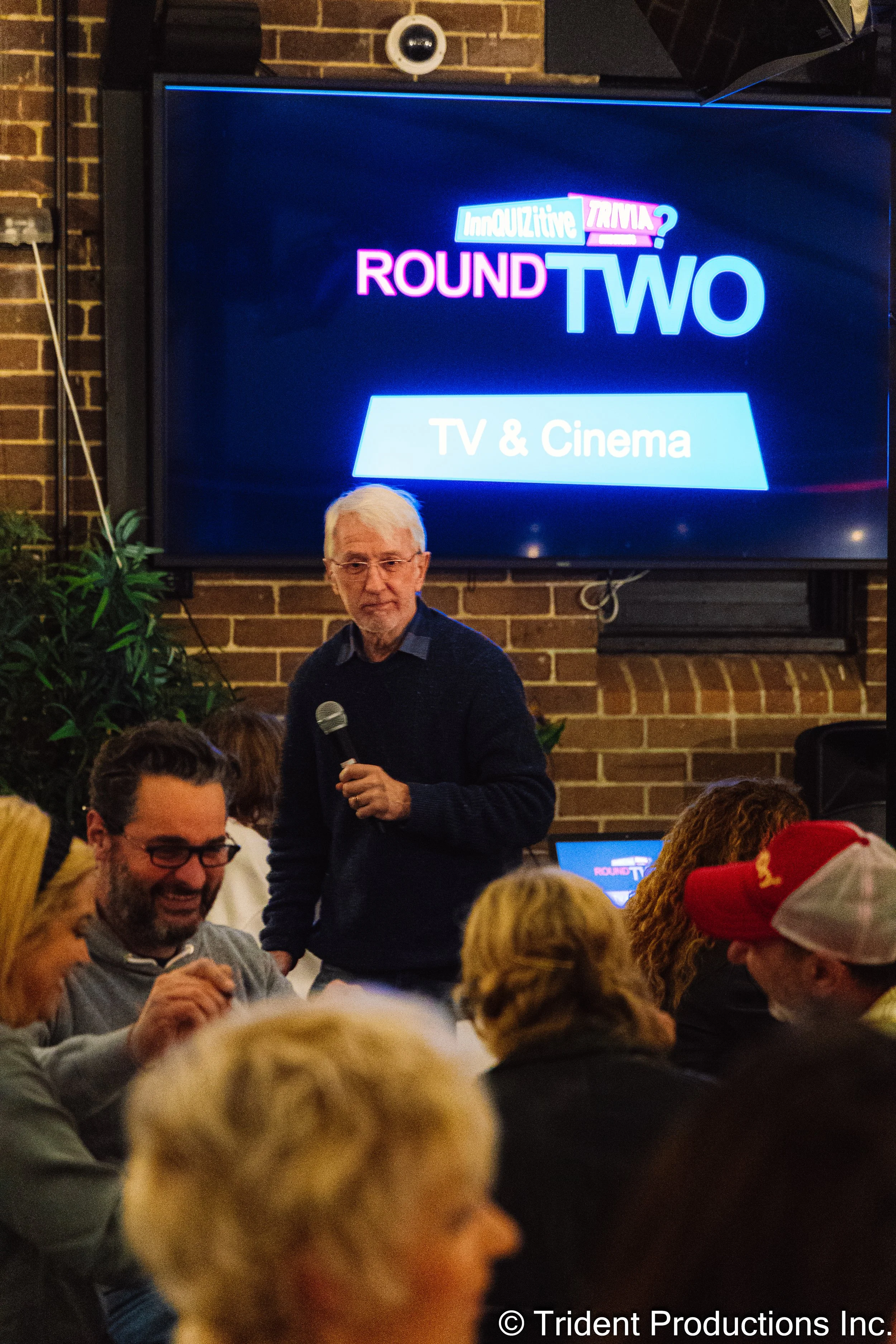 A man with white hair and glasses holding a microphone speaks to an audience at an event. Behind him on a large screen are the words 'Round Two TV & Cinema' and 'InnoQuizitive Trivia?' in colorful text.
