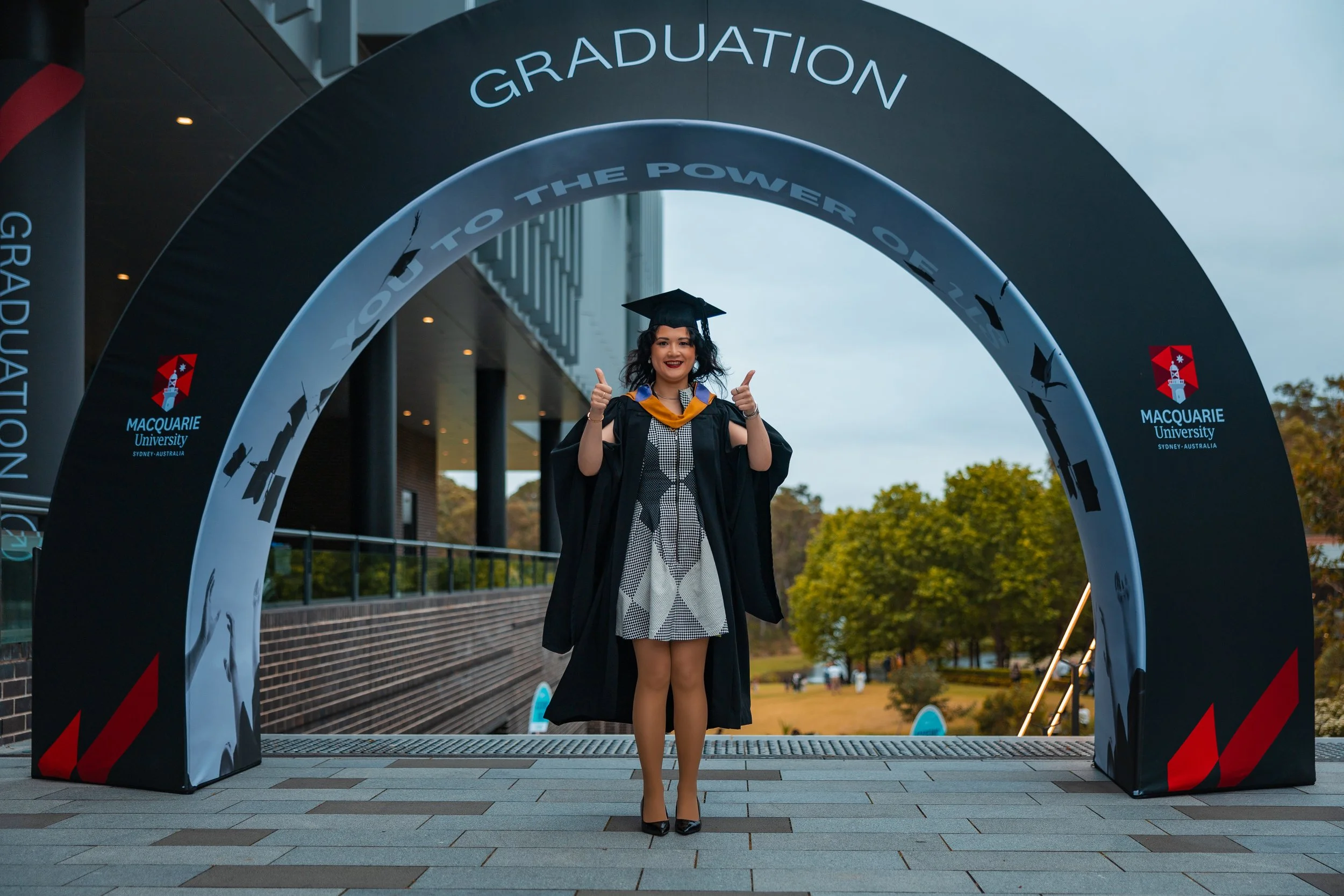 A woman in a graduation gown and cap standing under a graduation arch at Macquarie University, giving a thumbs-up and smiling.