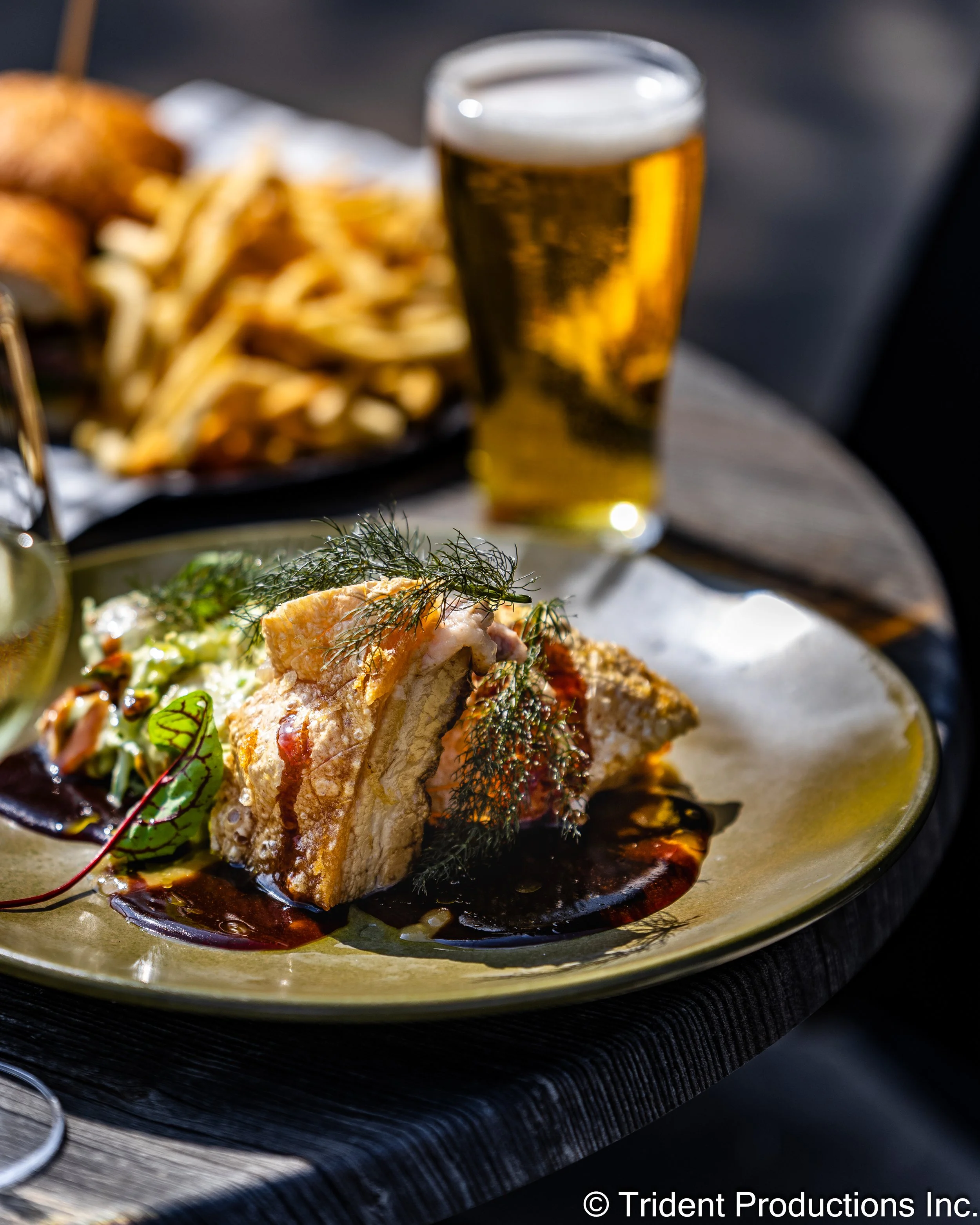 A plate with a piece of fish garnished with herbs and sauce, with a side of fries and a drink of beer in the background.