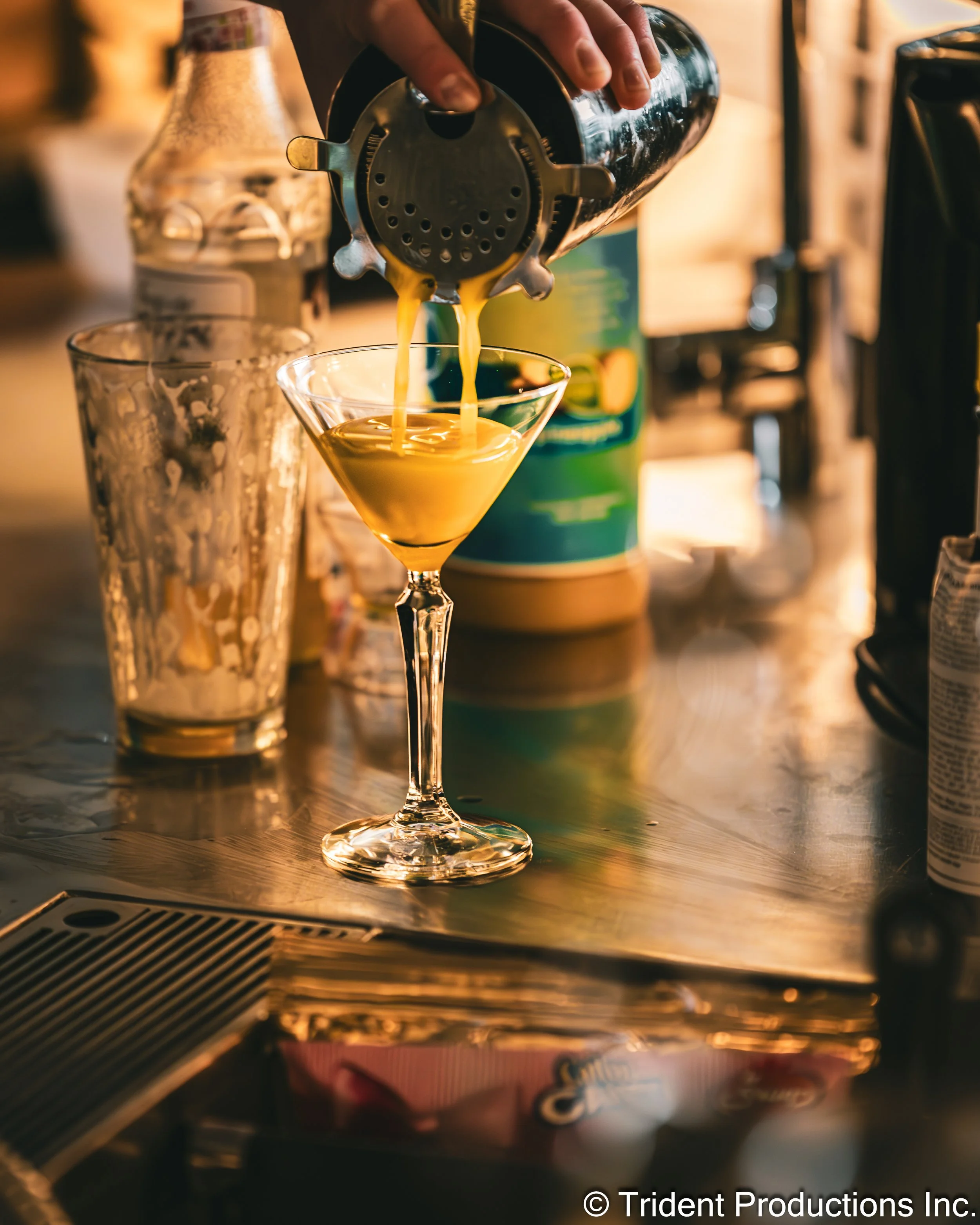 A person pouring orange juice from a cocktail shaker into a martini glass on a bar counter, with various bar supplies and bottles in the background.