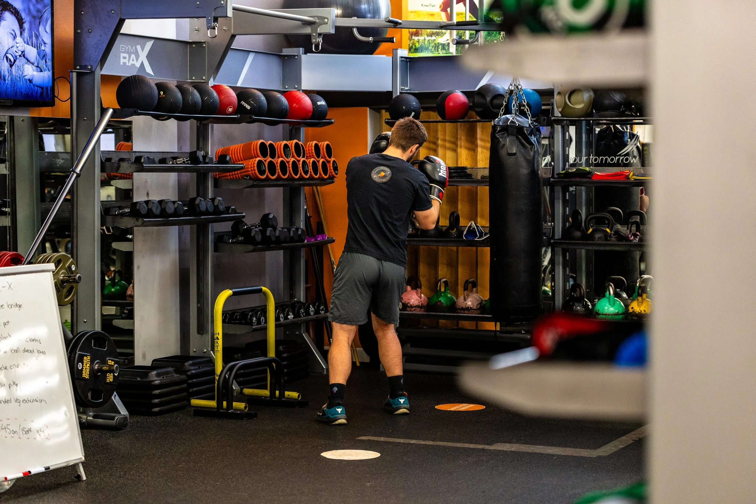 A man wearing black shorts, black t-shirt, and boxing gloves practicing punches near a black punching bag in a gym equipped with kettlebells, dumbbells, medicine balls, foam rollers, and a television.